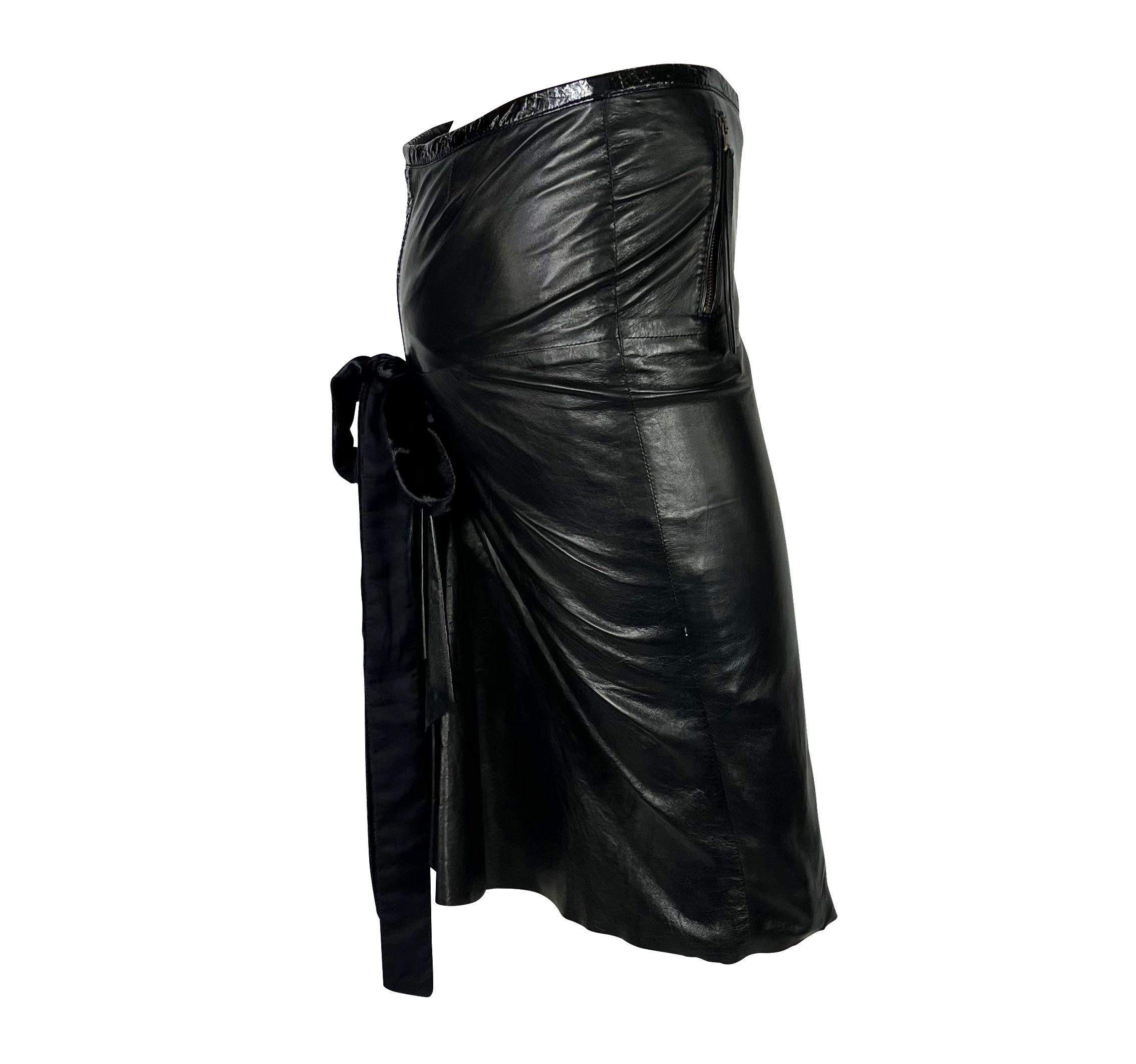 Presenting a black leather Roberto Cavalli pleated skirt. From the 2000s, this entirely leather skirt features a patent leather waistband, zipper pockets at the front, and ruching at the back. This skirt is complete with a velvet bow cascading down
