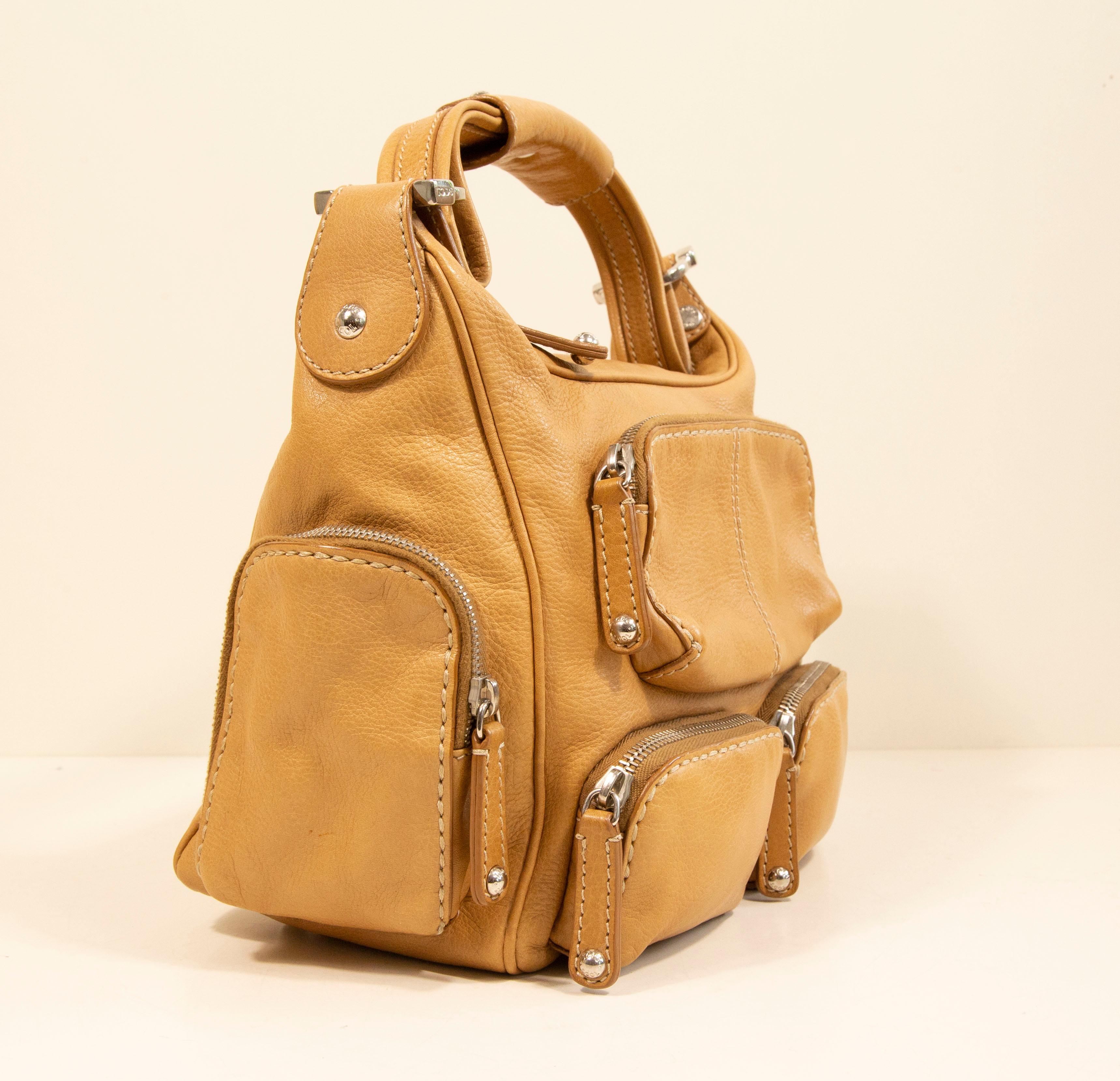 Tod's shoulder bag made of tan leather with silver toned hardware. The interior is lined with beige fabric, and next to the major compartment, it features one side pocket with a zipper. In addition, the bag has six zipped pockets on the outside. The