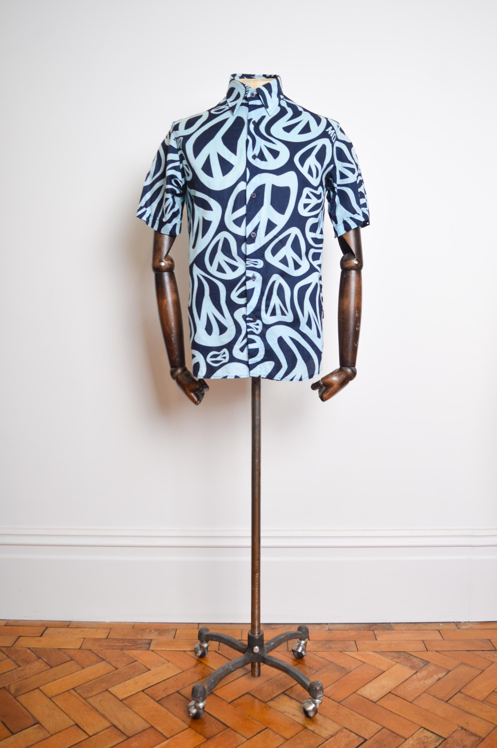 Witziges, kurzärmeliges Vintage 'MOSCHINO' Shirt aus bedruckter blauer Baumwolle mit wellenförmigem Peace-Symbol-Druck aus den frühen 2000er Jahren. 

GEFERTIGT IN ITALIEN.   

Merkmale: Knöpfe auf der Vorderseite, Kragen, kurze Ärmel, Brusttasche,