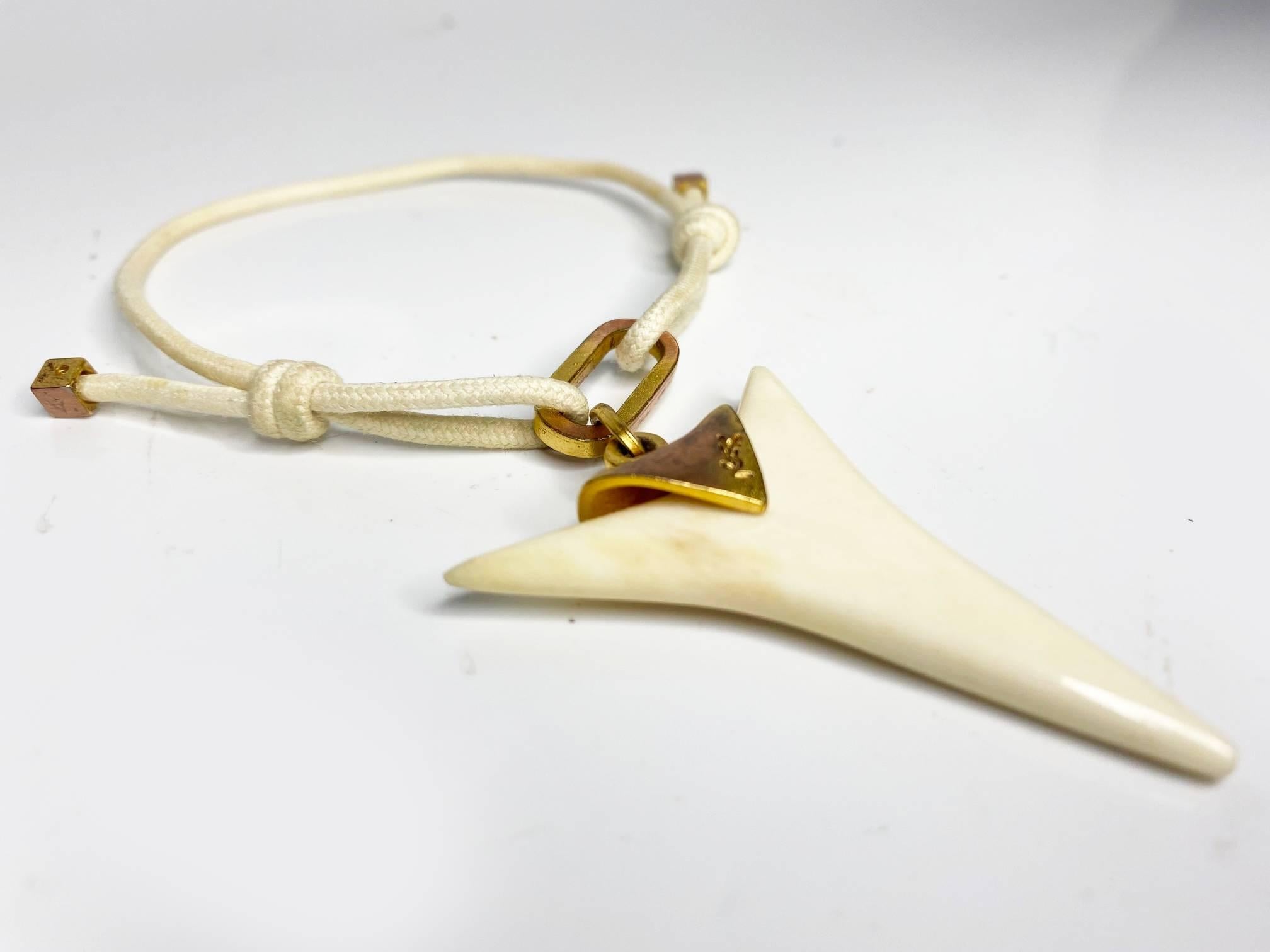 Yves Saint Laurent bracelet, shark shaped horn charm, logo on gold tone metalware, cream white cord bracelet. Made with a signature logo, this bracelet is sure to be a statement piece. Its luxurious and eye-catching materials bring a sense of