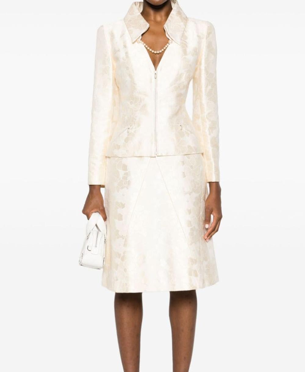 Tailleur jupe deux pièces à motif camélia ivoire clair Chanel, tissu mélangé coton-soie, motif Camélia signature, embellissement en fausses perles.
Pour la veste qui présente un col classique, une encolure en V, une fermeture à glissière