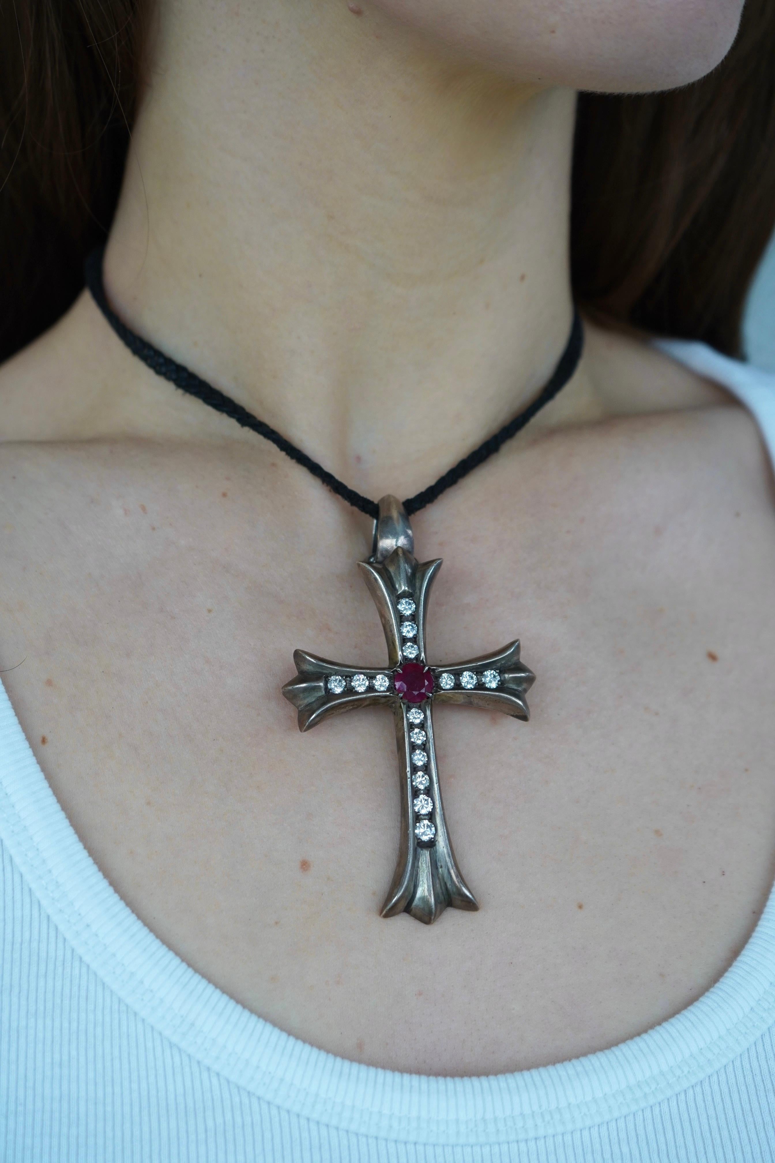 Este collar Chrome Hearts lleva un gran colgante en forma de cruz de plata de ley adornado con un rubí central y quince diamantes talla redonda, engastados uniformemente a lo largo de los cuatro brazos de la cruz. El diseño muestra el estilo gótico
