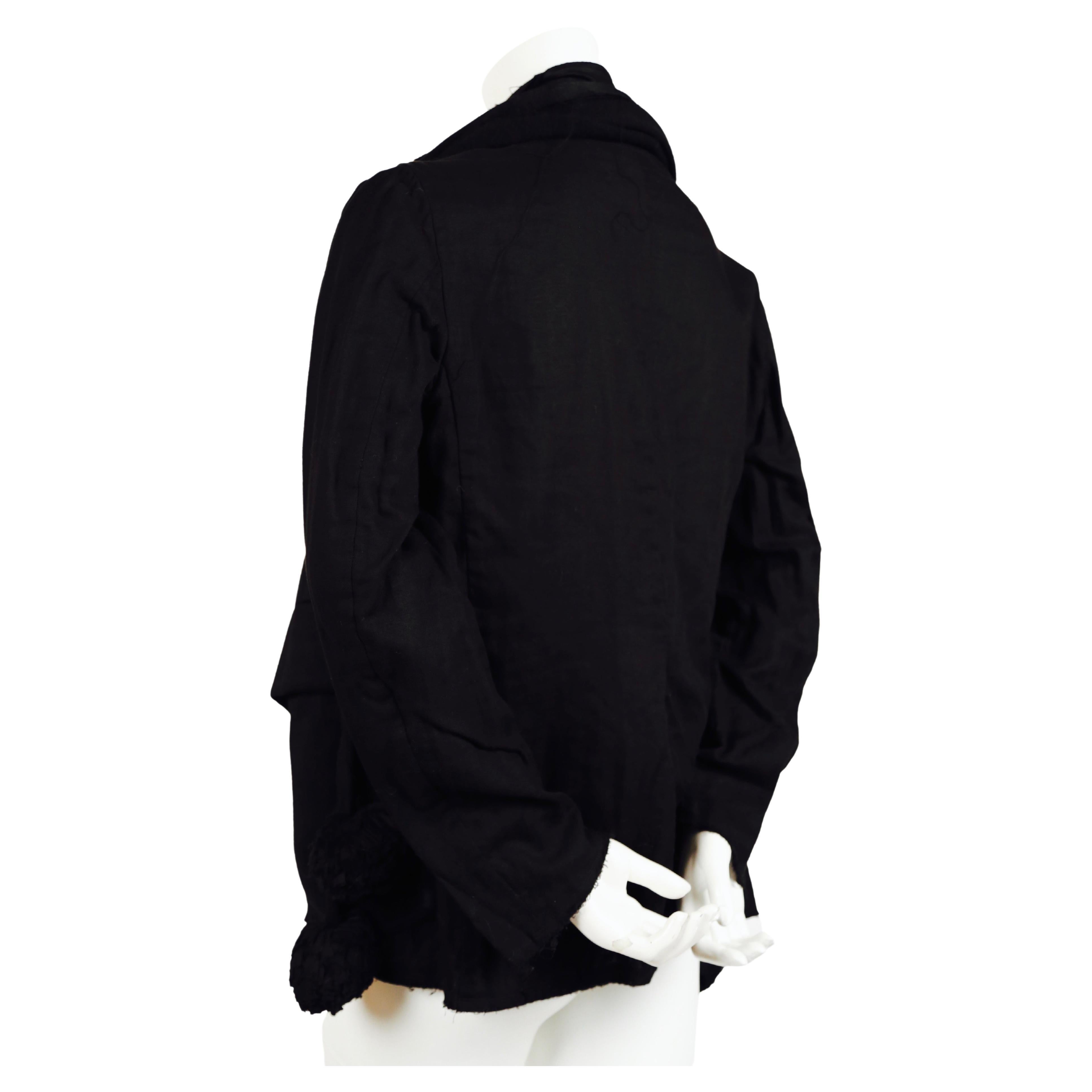 Black jacket with scarf overlay and pom pom details designed by Rei Kawakubo for Comme Des Garcons dating to spring of 2003. 'Scarf' can be worn completely over the shoulders/arms or gathered as shown. Jacket has a very unstructured fit. Raw edges