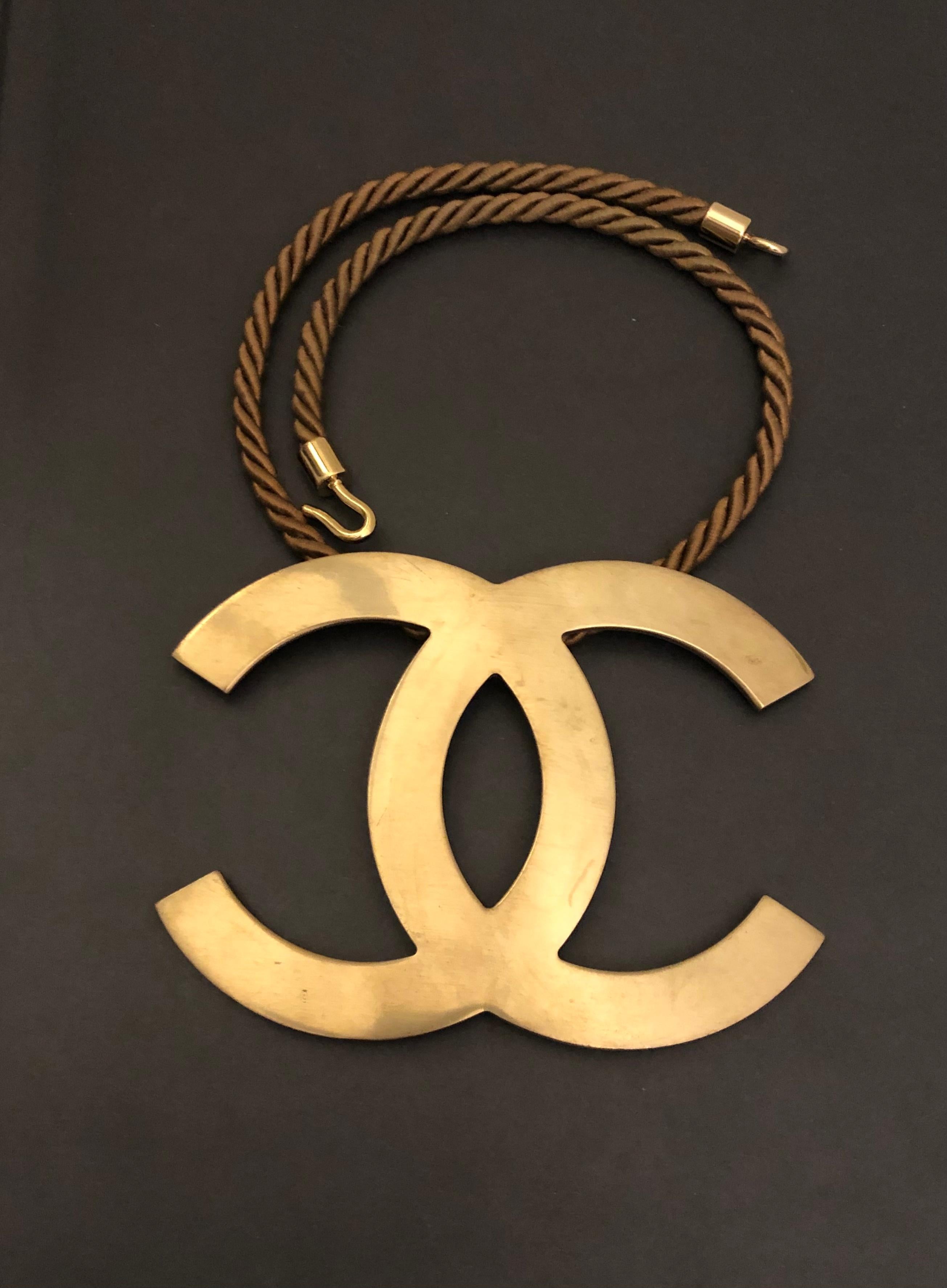 This CHANEL massive statement necklace is crafted of gold toned metal featuring a massive CC charm and silk rope in brown. Length measures approximately 40 cm Charm 10 x 8 cm. Stamped Chanel 05P made in France. 

Condition - Excellent with minimal