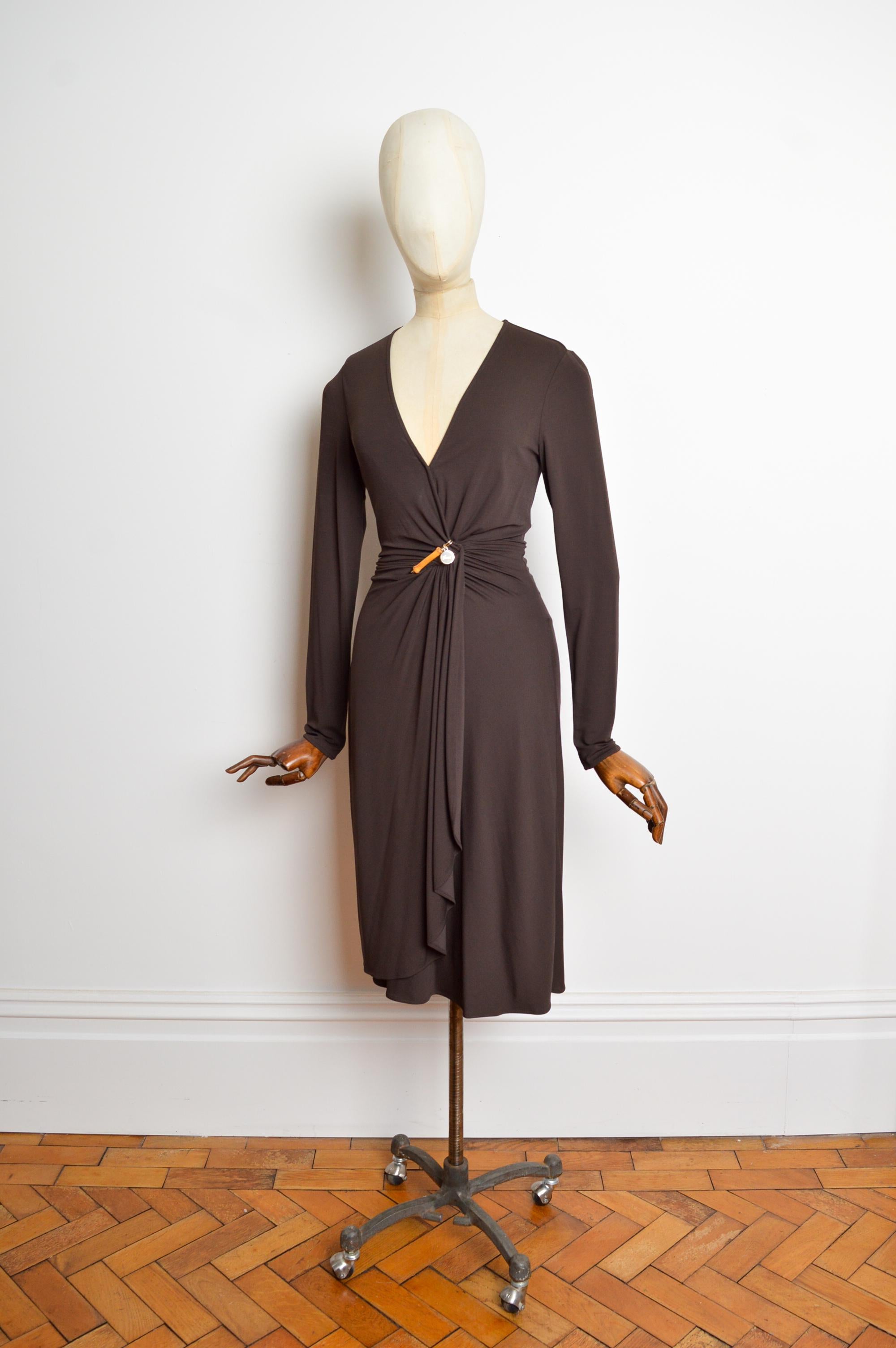 Robe intemporelle GUCCI 2006 de Frida Giannini, réalisée dans une matière épaisse et extensible marron chocolat, avec un pendentif GUCCI en bambou entre le buste, des manches longues, une taille ajustée et un ourlet jusqu'au genou. 
La robe est