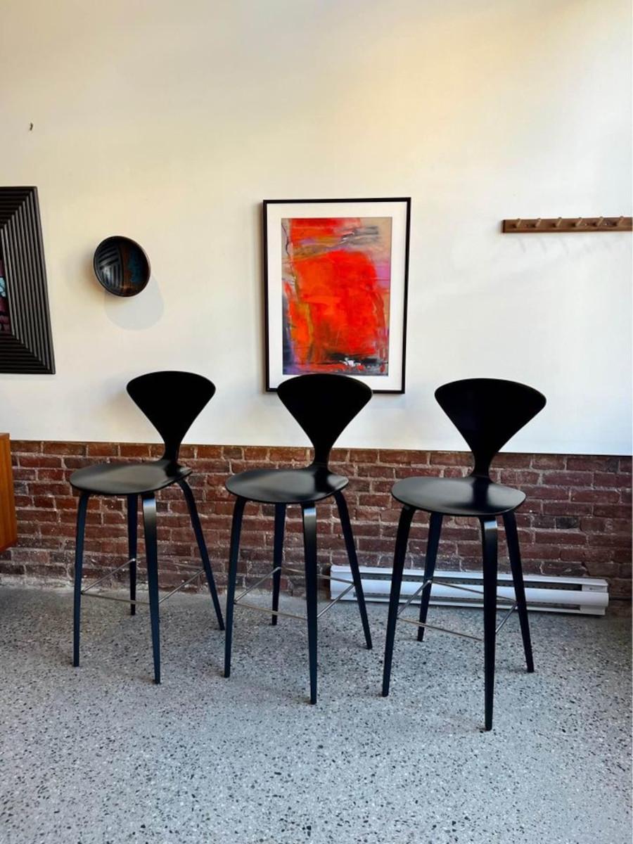 Presenting three 2008 black Cherner barstools, featuring a sophisticated wood base and chrome footrest. Reviving Norman Cherner's iconic designs, the Cherner Chair Company meticulously crafts these pieces with original drawings and specifications,