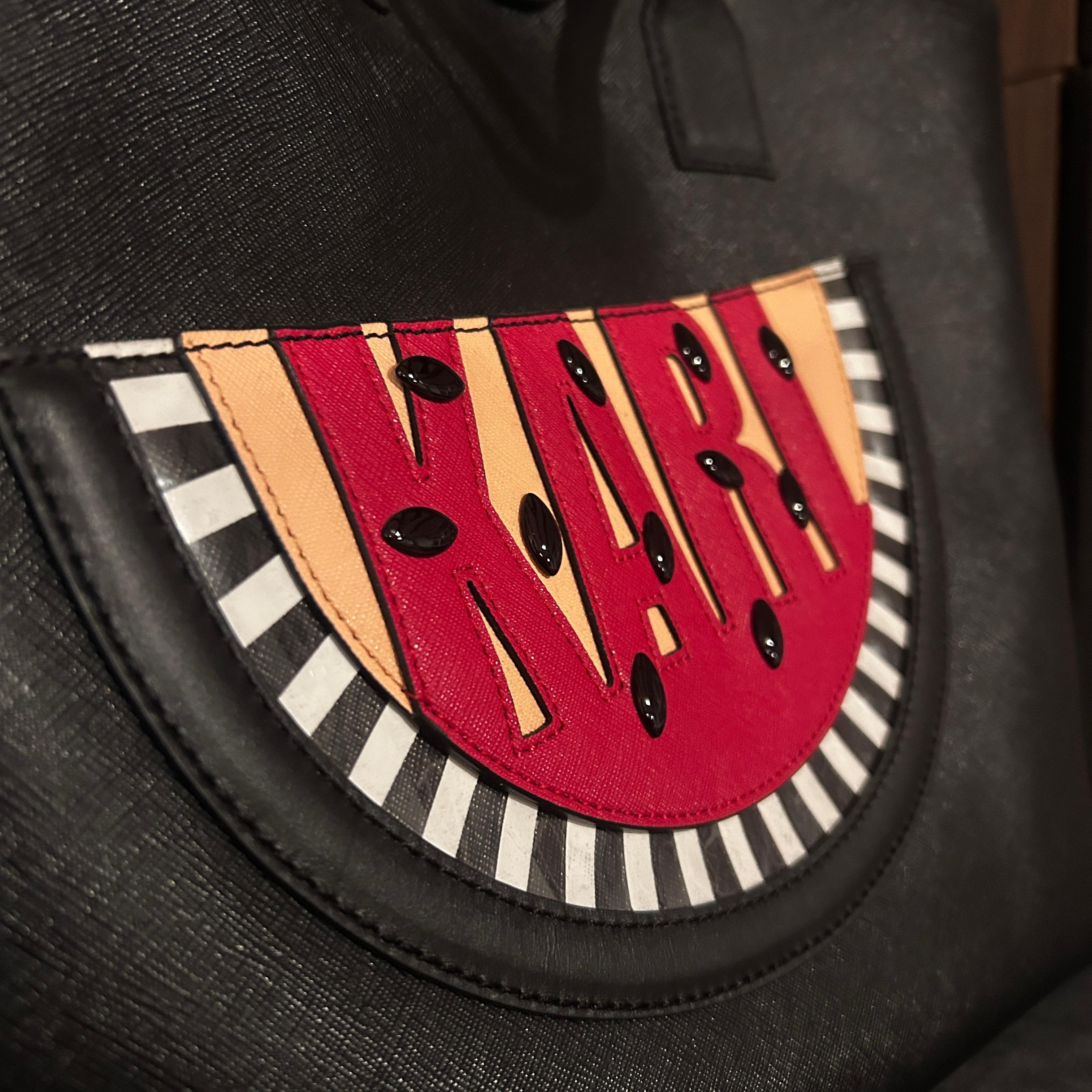 This Karl Lagerfeld bag is a large black tote with a striking and playful design on the front. The central feature is a semi-circle graphic resembling a watermelon slice, with black seeds and vibrant red, orange, and pink colors. The word 