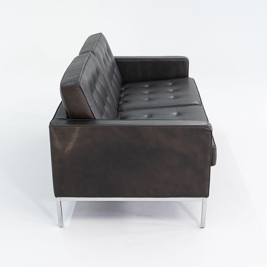 This is a two-seater Florence Knoll settee sofa, model 1205S2, originally designed by Florence Knoll in 1954. This example dates to 2013, and is upholstered in black leather. The design features a square tufted back and seat, and it sits atop a