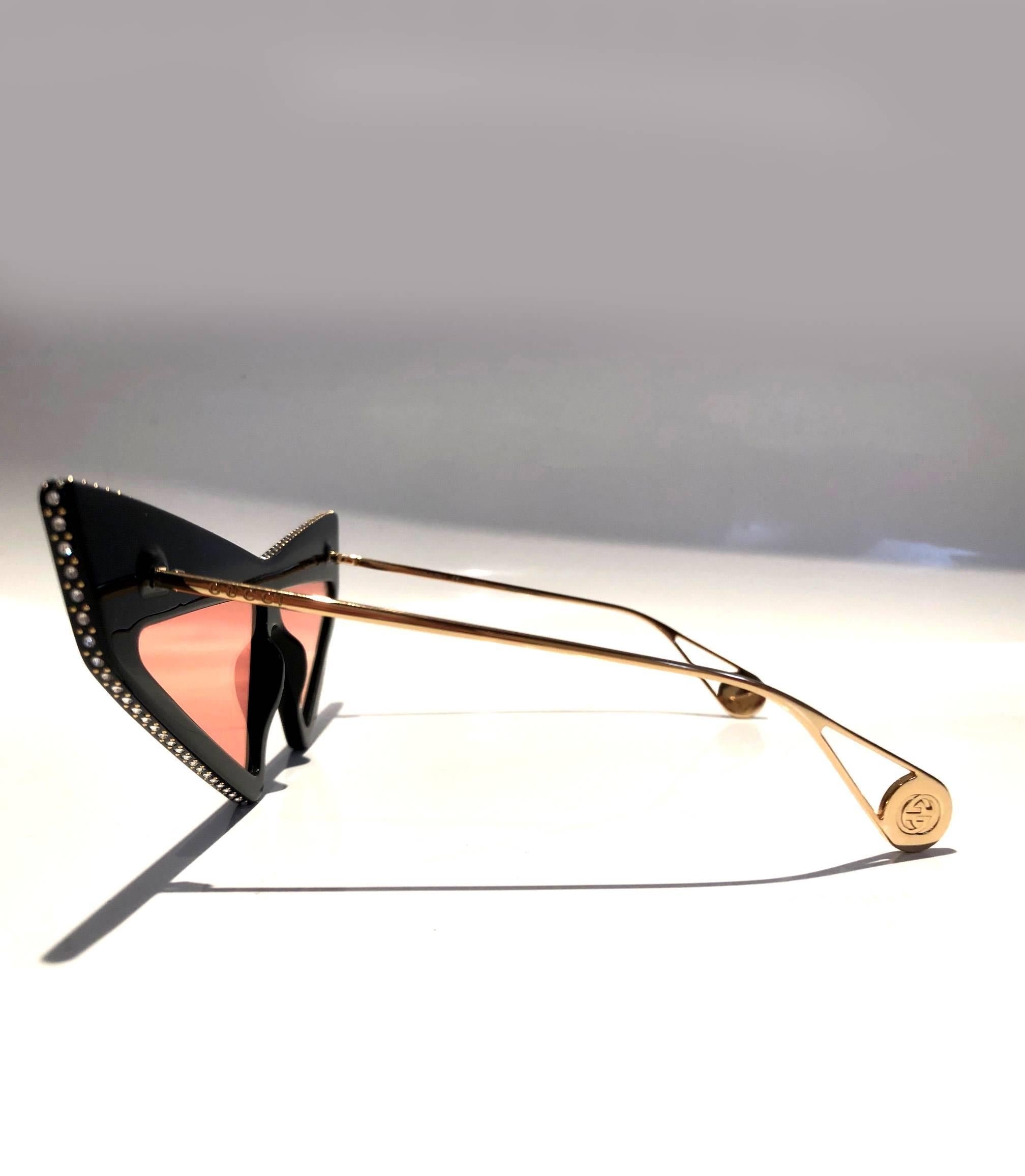 Gucci GG0430S 001 70 sunglasses feature an over exaggerated geometric cat eye frame, black frame, orange/pink lenses, these premium Havana acetate sunglasses are embellished with crystal Swarovski on the profile and feature stylish gold coated arms.
