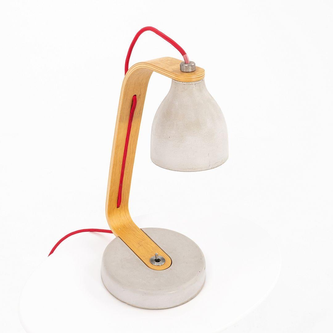 This is a Heavy Desk light lamp, designed by Benjamin Hubert for Decode London in 2009. This particular example dates to 2019. The piece is made from thin-walled cast concrete, and has an arm of oak veneer. The cord has been wrapped with red