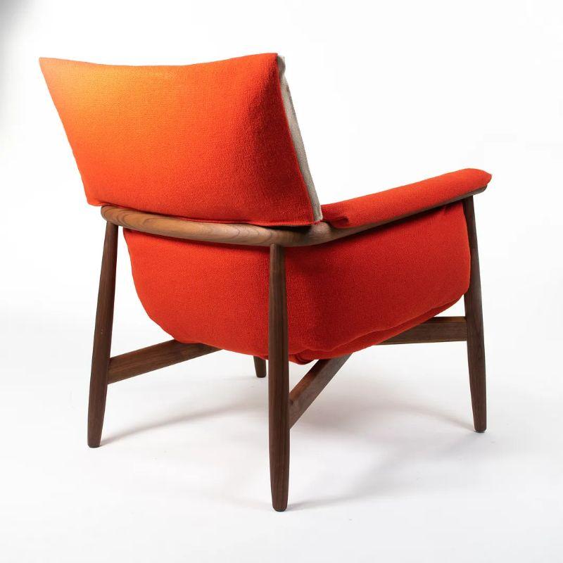This is an EO15 Embrace Lounge Chair with a solid walnut frame, red / orange fabric upholstery, and natural edge strip. The chair was designed by EOOS and produced by Carl Hansen & Son in Denmark. The chair dates to circa 2020 and is guaranteed as