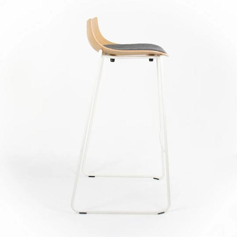 This is a BA004S Preludia Bar Stool, designed by Brad Ascalon and produced by Carl Hansen & Son in Denmark. The stool is made with a white powder-coated steel frame and a gray fabric seat with oak base. The stool dates to circa 2021 and guaranteed