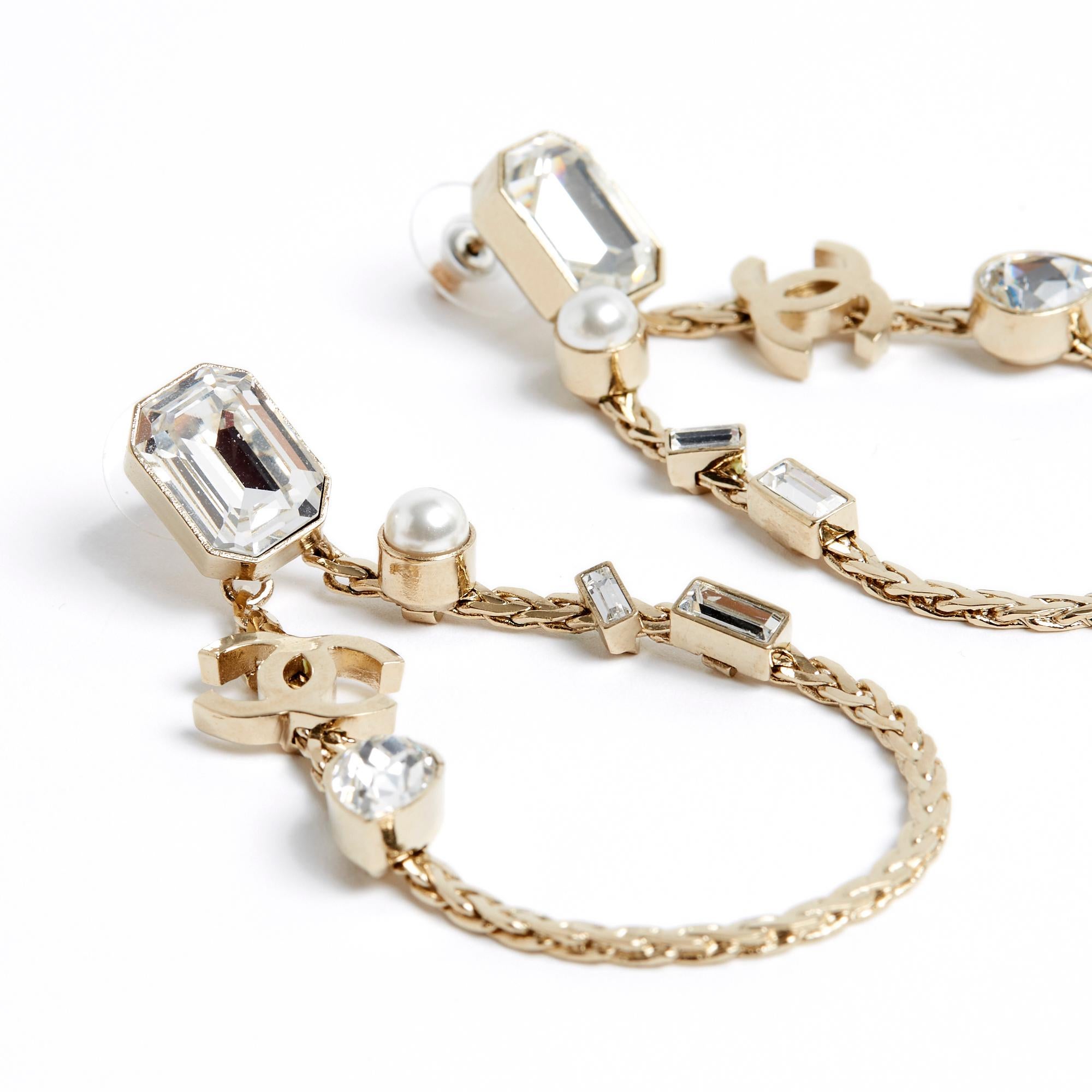 Chanel earrings from the SS2022 collection in lightly golden metal, composed of a stud encrusted with a large emerald-cut white rhinestone and a large flat chain buckle inlaid with rhinestones and fancy pearl and a CC symbol. Total height of the