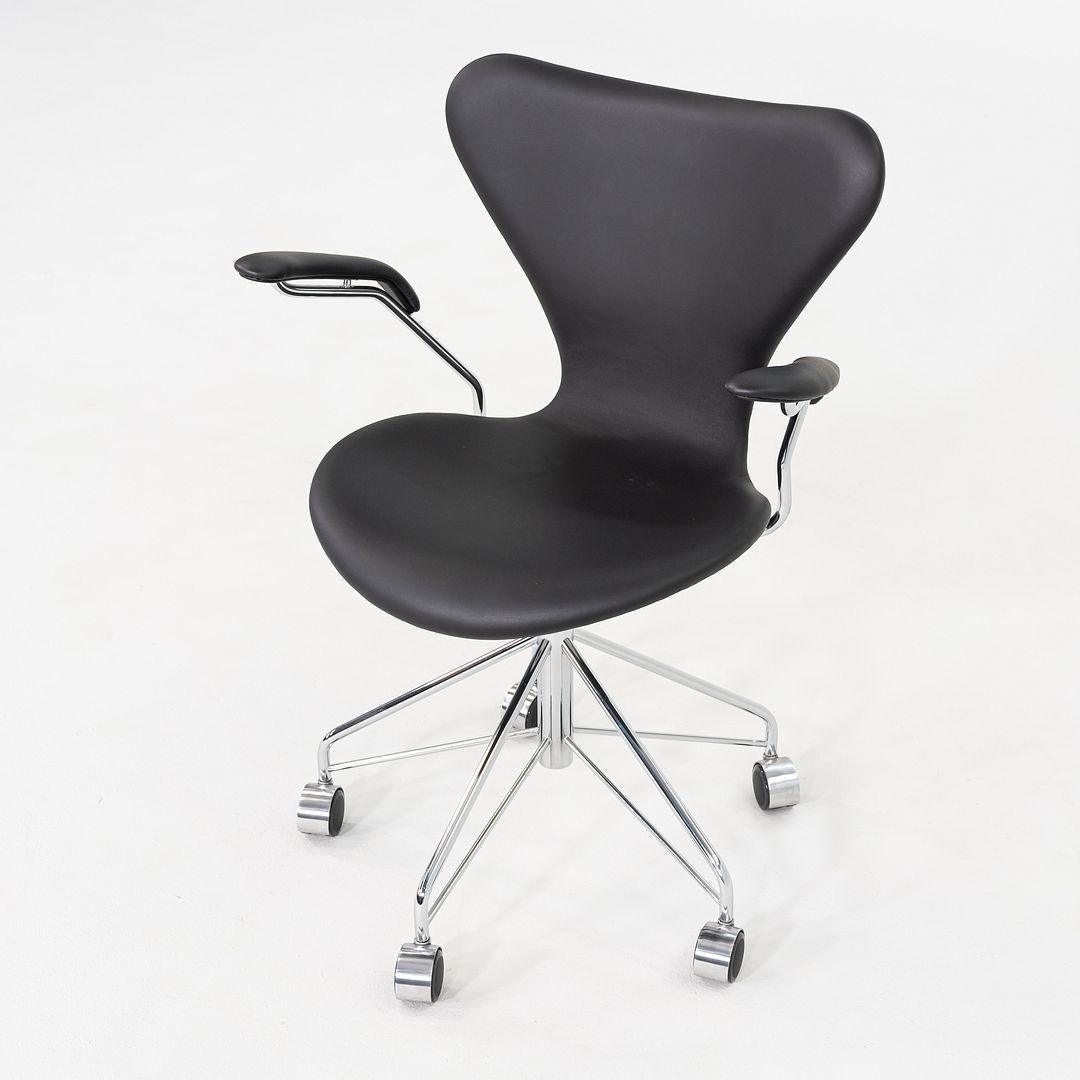 This is a Series 7 Desk Chair with Arms, originally designed by Arne Jacobsen in 1970. This example was produced by Fritz Hansen in Denmark in 2023. The chair features a molded plywood shell upholstered in black leather, paired with slender chrome