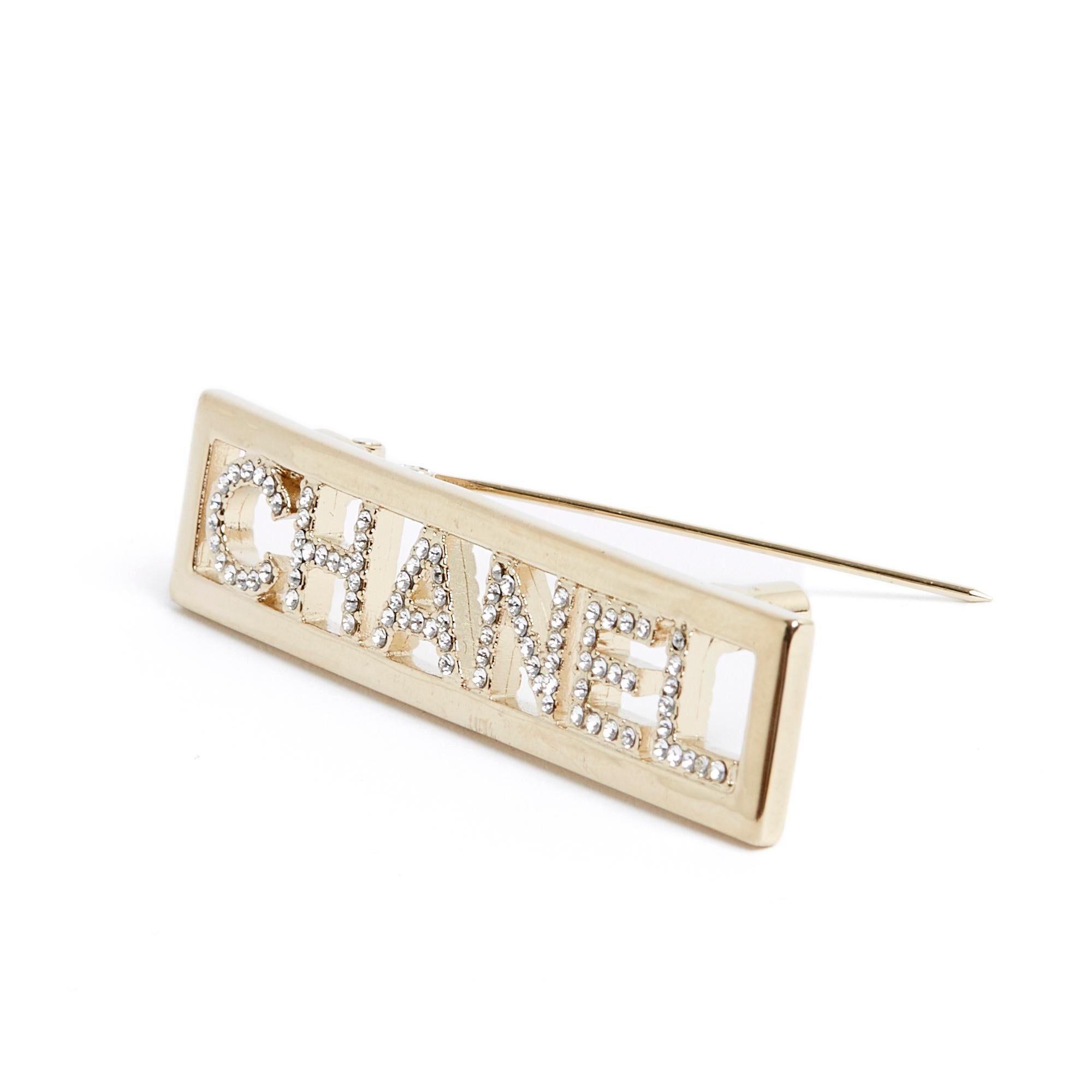 Chanel brooch in gold metal decorated with the CHANEL inscription encrusted with white rhinestones. Width 6.2 cm x height 1.5 cm. The brooch is sold without invoice or original packaging but it is in very good condition, from a very recent