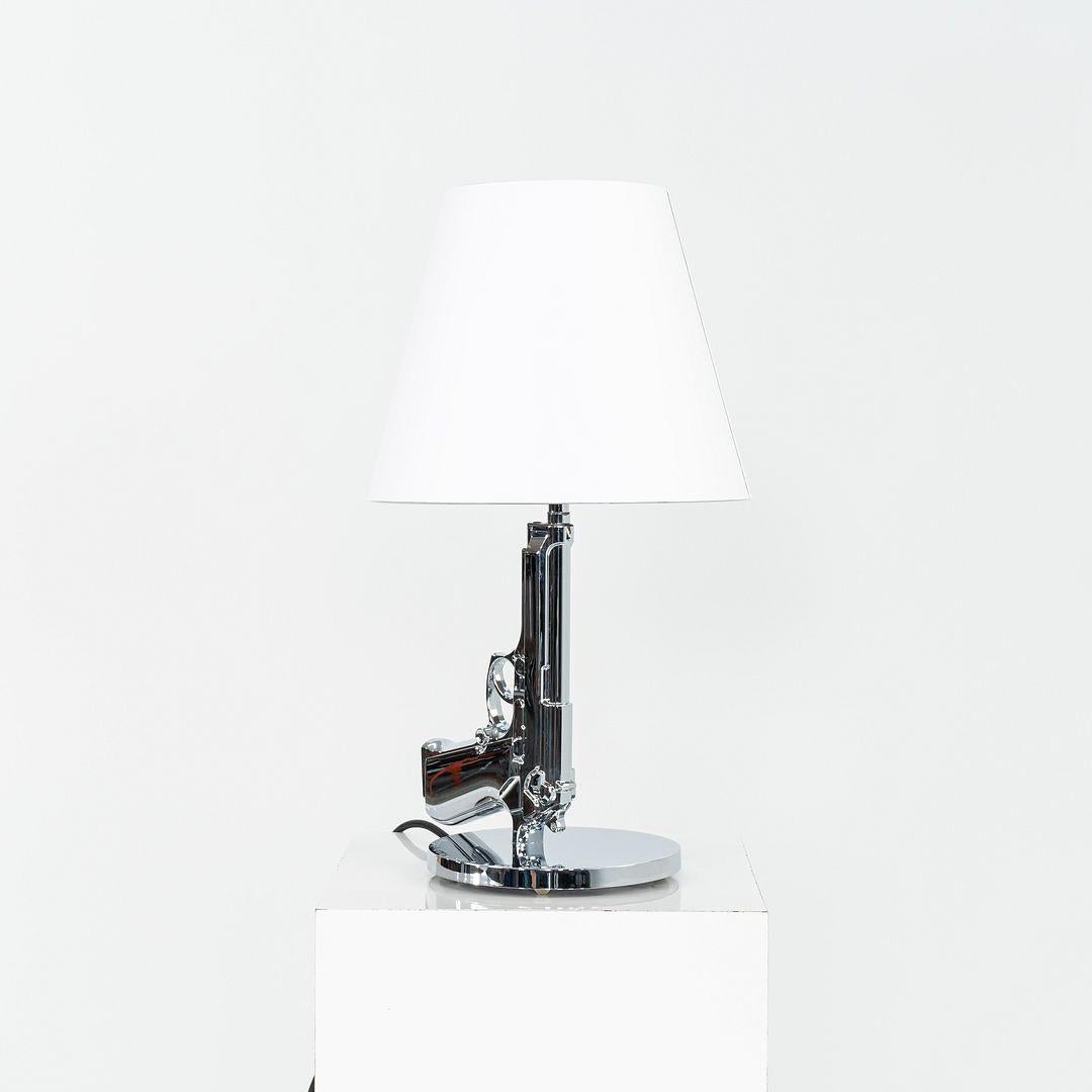 This is a Gun Bedside Lamp, originally designed by Phillippe Starck in 2005. This particular example was produced by Flos in Italy C. 2023. The lamp is comprised of an aluminum base with a paper shade. 
 
 The lamp measures 9.29 inches at the widest