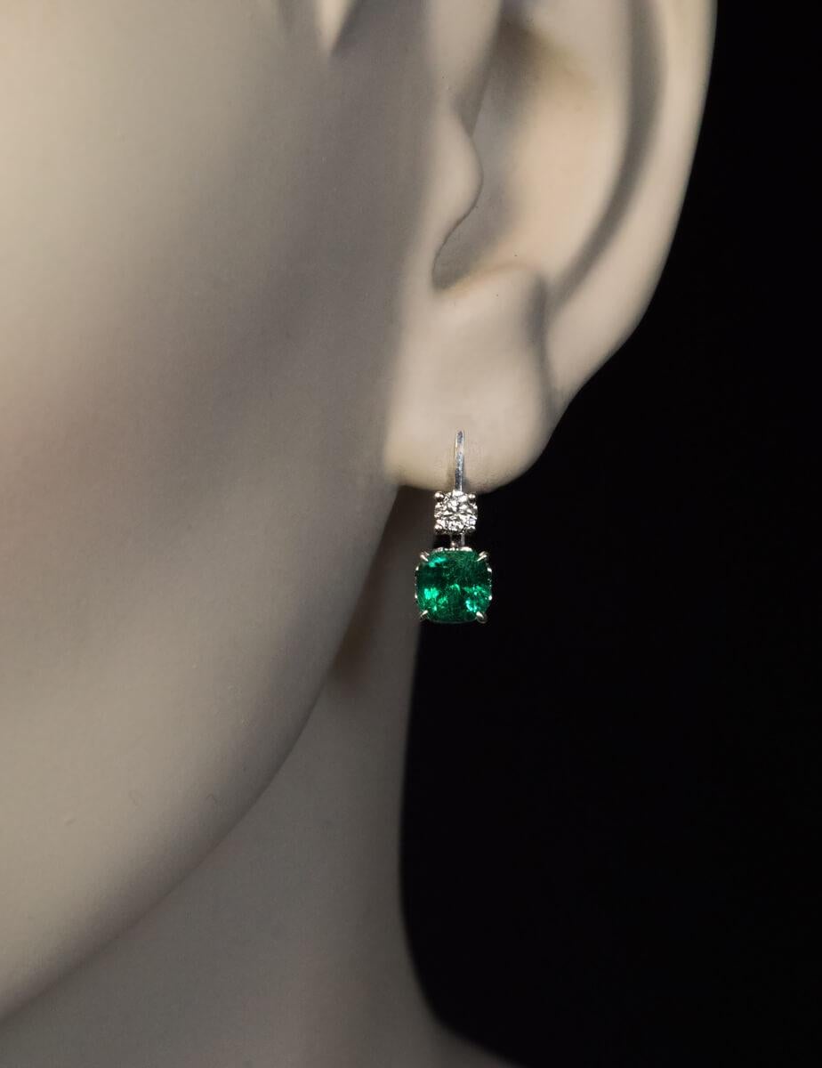 These modern 14K white gold earrings feature two cushion cut Zambian emeralds of excellent vivid bluish green color and strong saturation. The emeralds (6 x 5.9 mm and 6 x 6 mm) are accented by two bright white brilliant cut diamonds (F-G color,