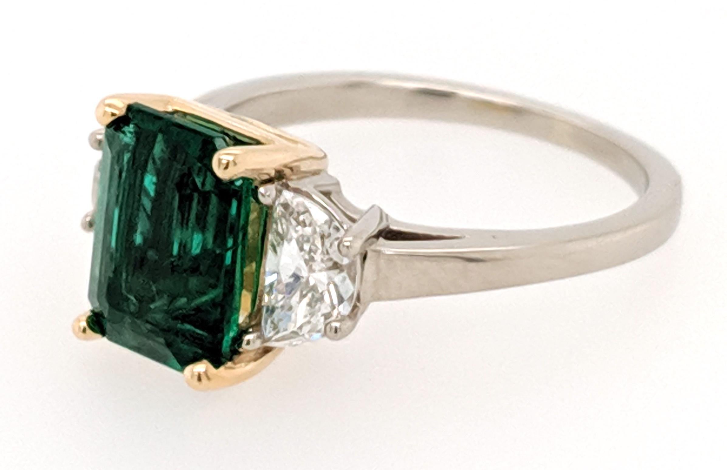The Emerald and Diamond 3 stone ring is crafted in 18k white gold with a 18k yellow gold box and features (1) Emerald cut Zambian Emerald weighing 2.05ct and flanked by (2) half moon diamonds weighing .63 cttw with a color of F/G and a clarity of