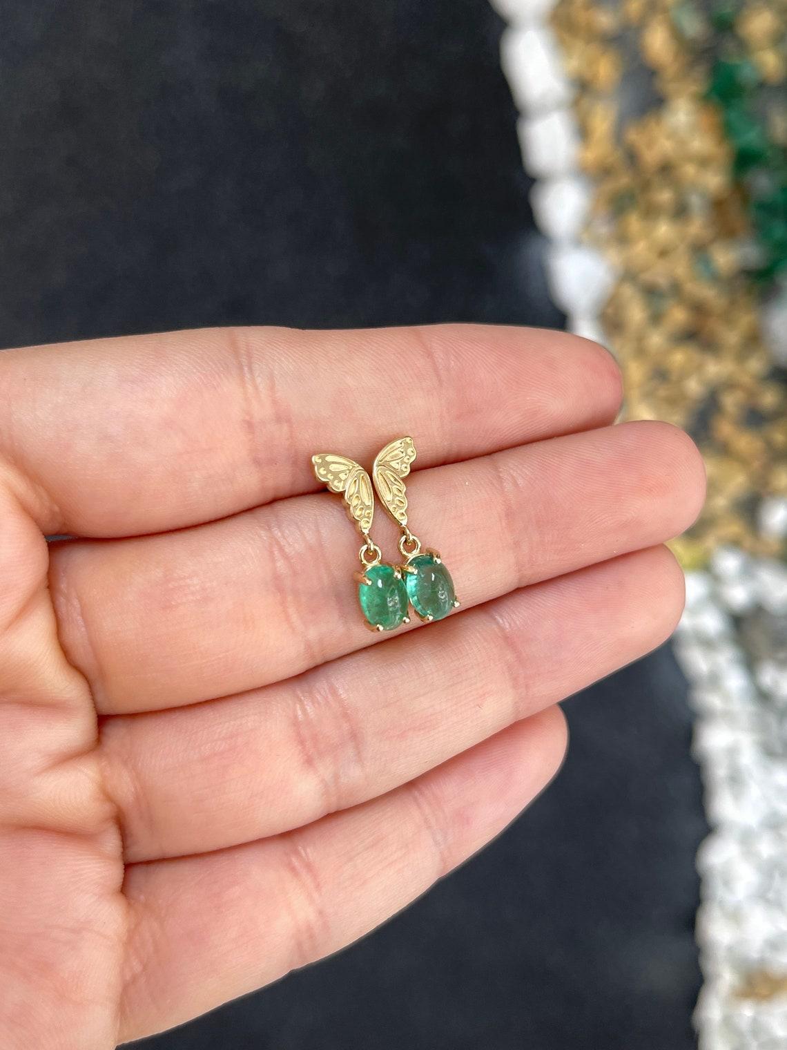 Aggiungi un tocco di stravagante eleganza al tuo ensemble con questi accattivanti orecchini con farfalla smeraldo. Ogni orecchino mostra la metà di una farfalla d'oro, ornata da smeraldi cabochon di taglio ovale che emanano un bellissimo colore