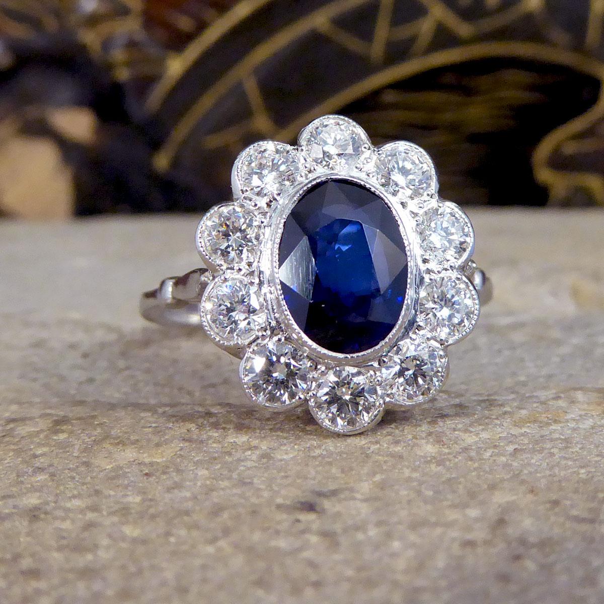 This contemporary ring holds a 2.07ct deep blue Sapphire in the centre of the ring and surrounded by 10 Modern Brilliant cut bright and clear Diamonds making a cluster. Complimenting the Sapphire gemstone is a Platinum rub over collar setting,