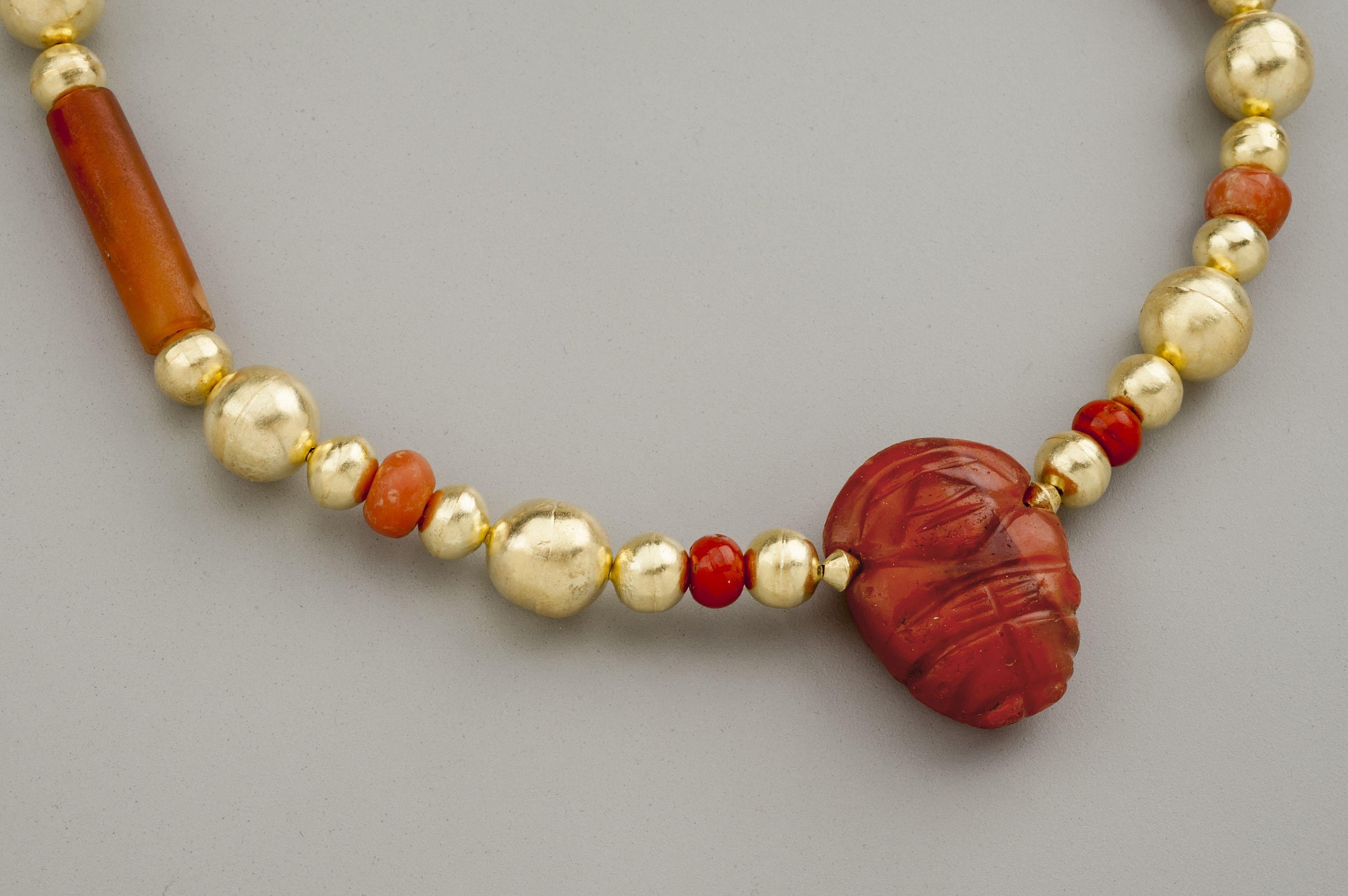 Twenty-eight round gold beads with ten carnelian beads, eight of which are flattened spheres and two are cylindrical tubes, with a carnelian bean shaped effigy of a of a man, presumably a chief. The stone pieces are said to be from the Tairona