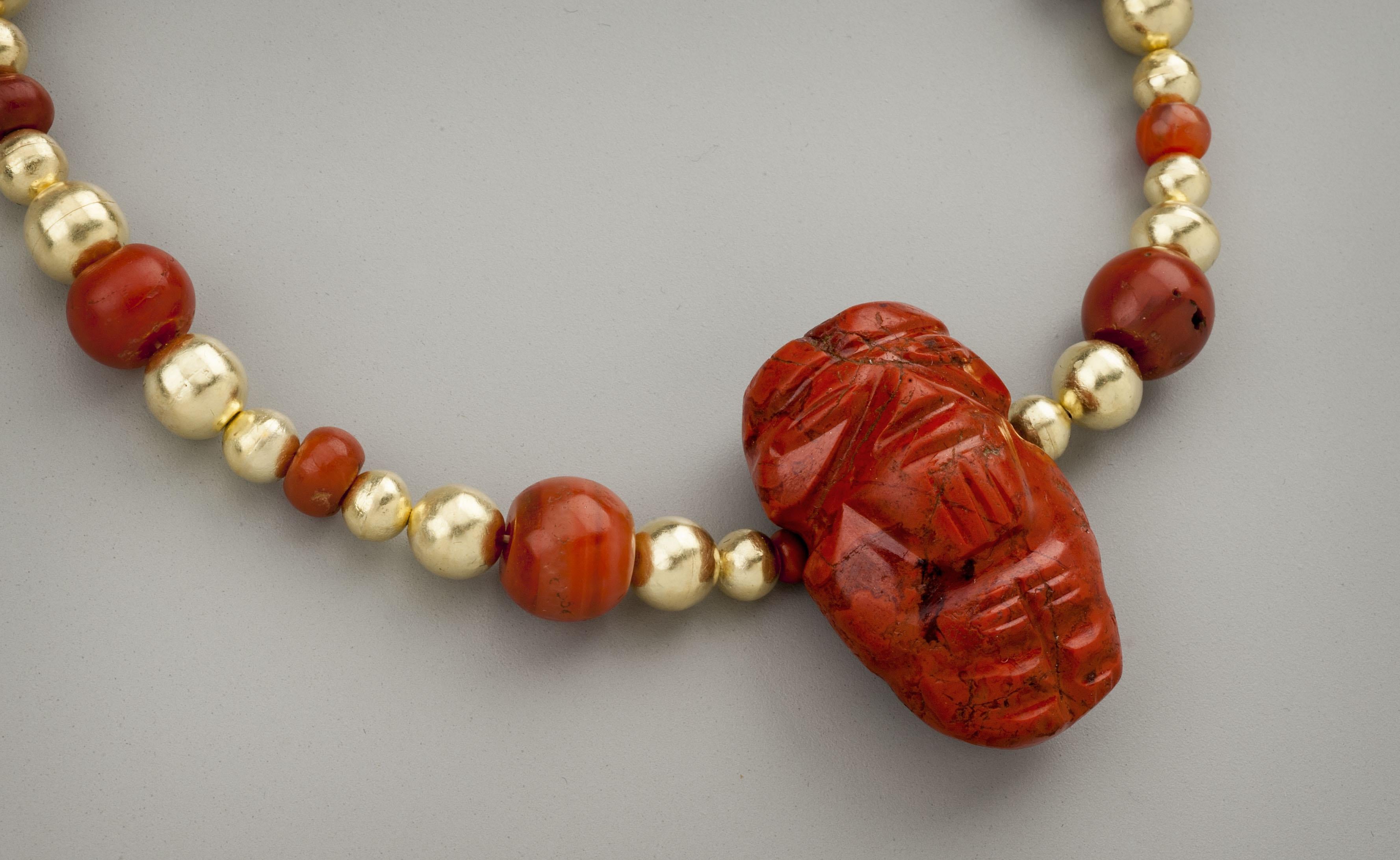 Twenty-six round gold beads with sixteen round carnelian beads and a carved jasper figure of a man pendant. The man, presumably a chief, holds his hands touching in front of his chest. A beard is indicated with three vertical marks. The eyes appear