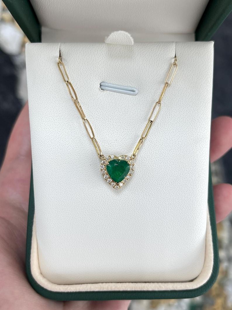 Questo squisito ciondolo presenta un affascinante smeraldo di terra tagliato a cuore dal colore verde intenso, splendidamente circondato da un'abbagliante aureola di diamanti. Il raro smeraldo è incastonato in una sicura montatura a tre punte,