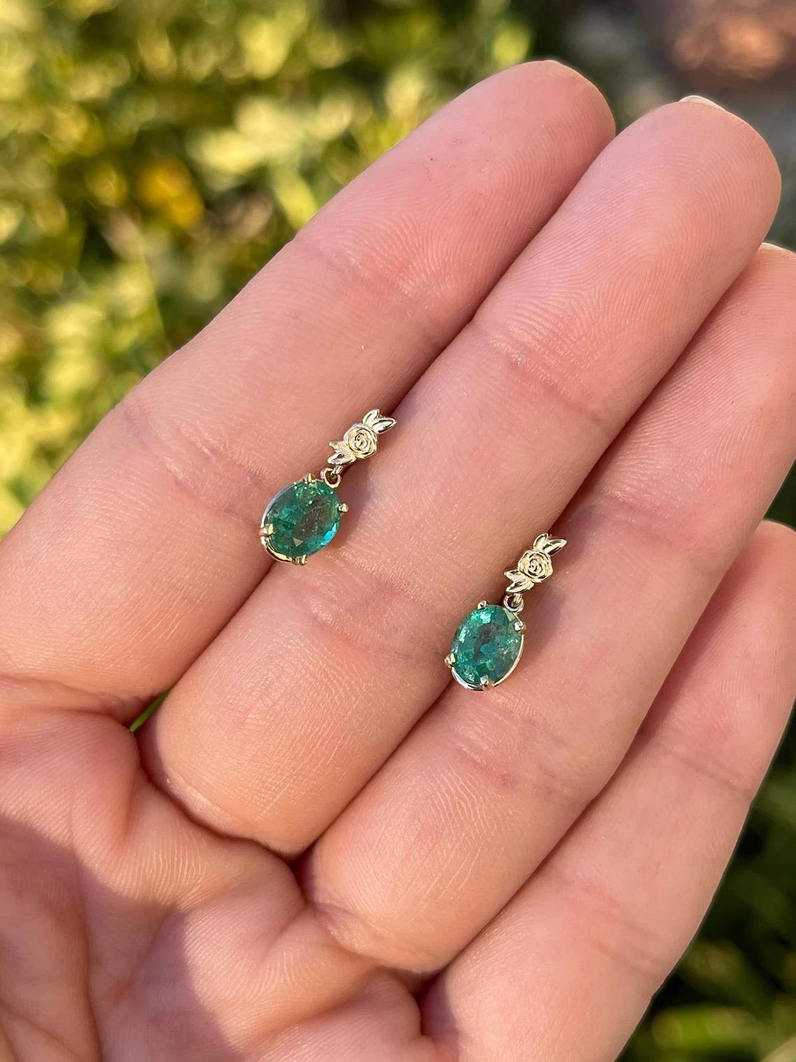 Displayed is a stunning pair of emerald and diamond, dangle earrings. Two gorgeous Zambian emerald-oval cuts totaling a full 2.0 carats, accented by rose golden details in a drop & dangle setting. The emeralds are prong set, allowing for the