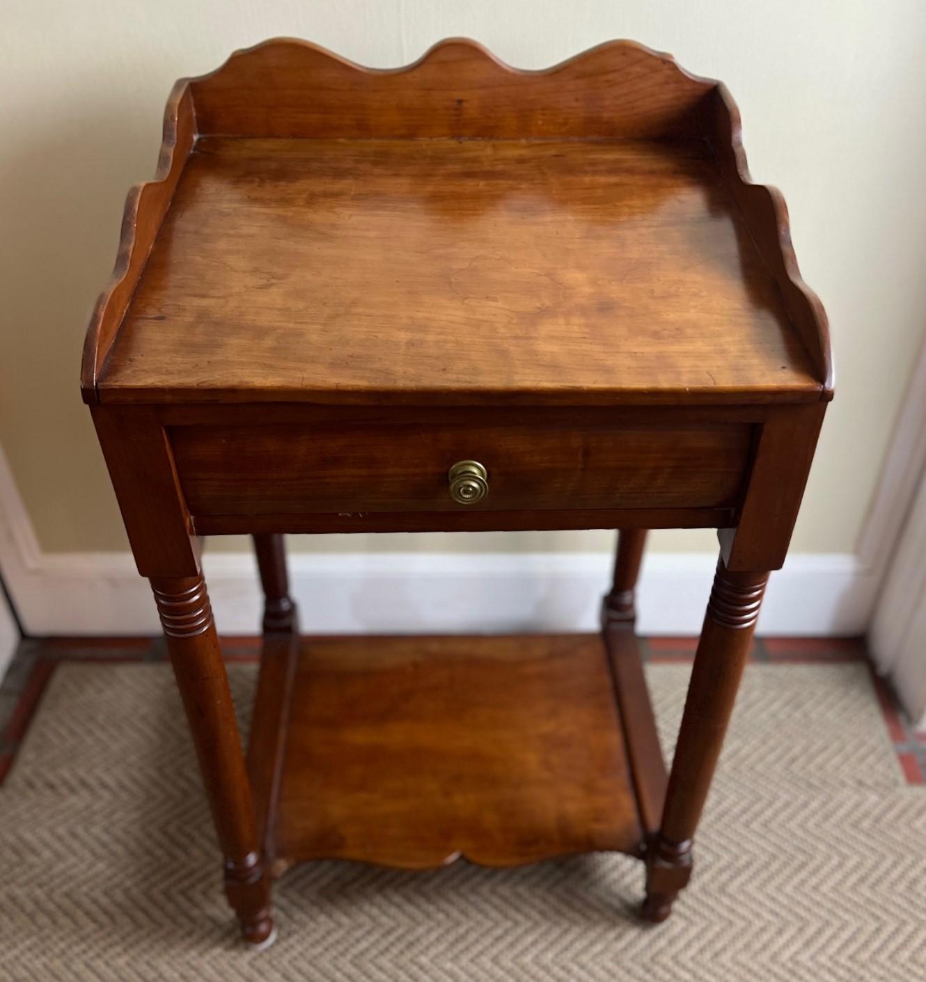 Late 20th C. rich honey colored wooden bedside table/nightstand with a drawer, low shelf and bobbin style turned legs. The table has nice details with scalloped gallery sides on the table top, a bow drawer front and a nicely curved lower shelf. This