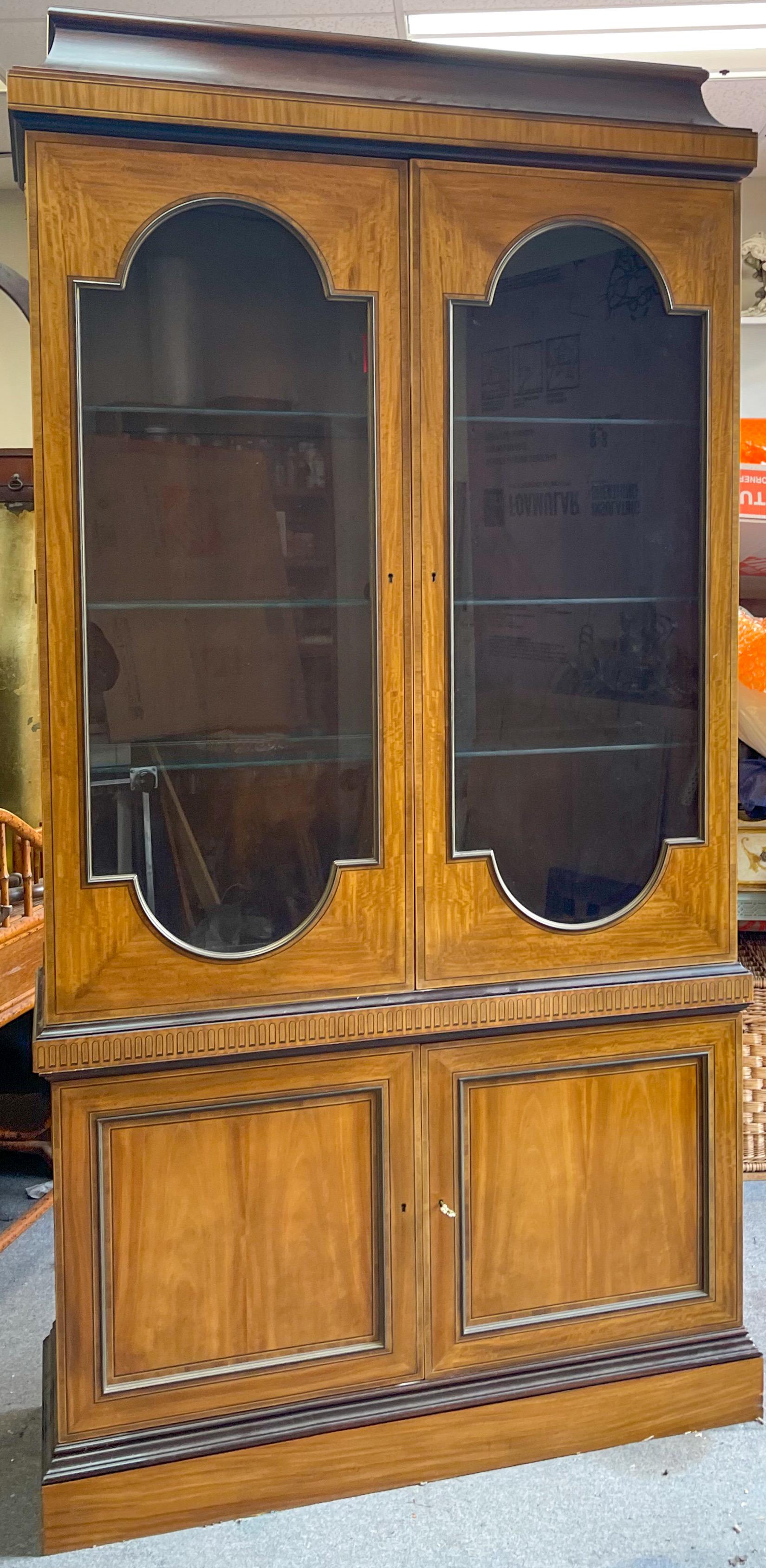This is a good looking cabinet! This is a neo-classical style cabinet by Baker Furniture Company. The top portion is lit, and it is two pieces. The cabinet is marked and in very good condition. It also includes the key.