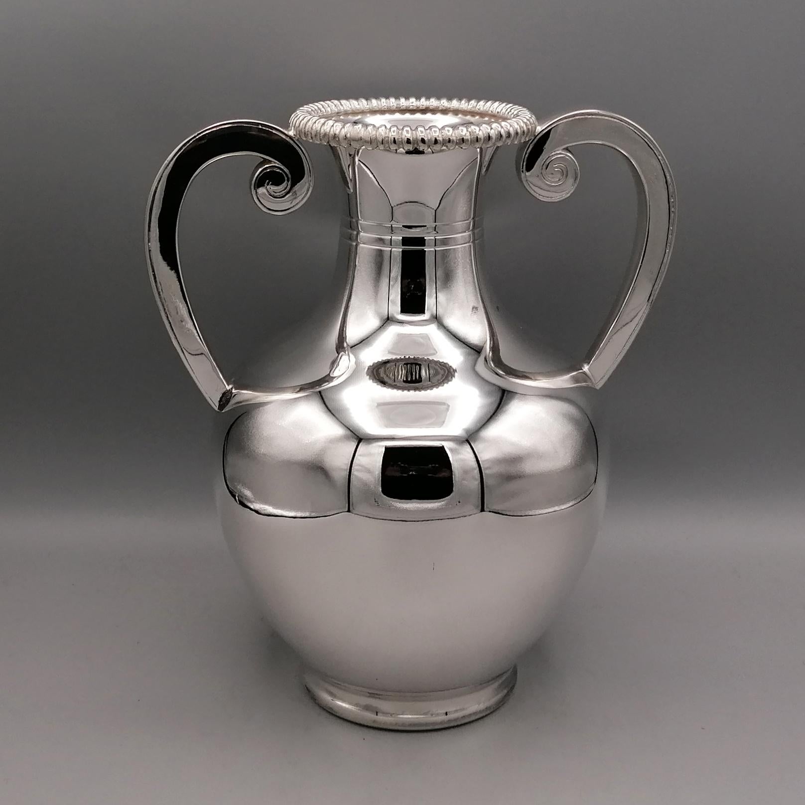 Amphora Vase mit Henkeln aus 800 massivem Silber im neoklassischen Stil.
Der Körper ist rund, abgerundet und völlig glatt. Nur am Hals der Amphora wurden 3 Rillen angebracht, um die Gleichförmigkeit der Struktur aufzubrechen.
Auf die Mündung der