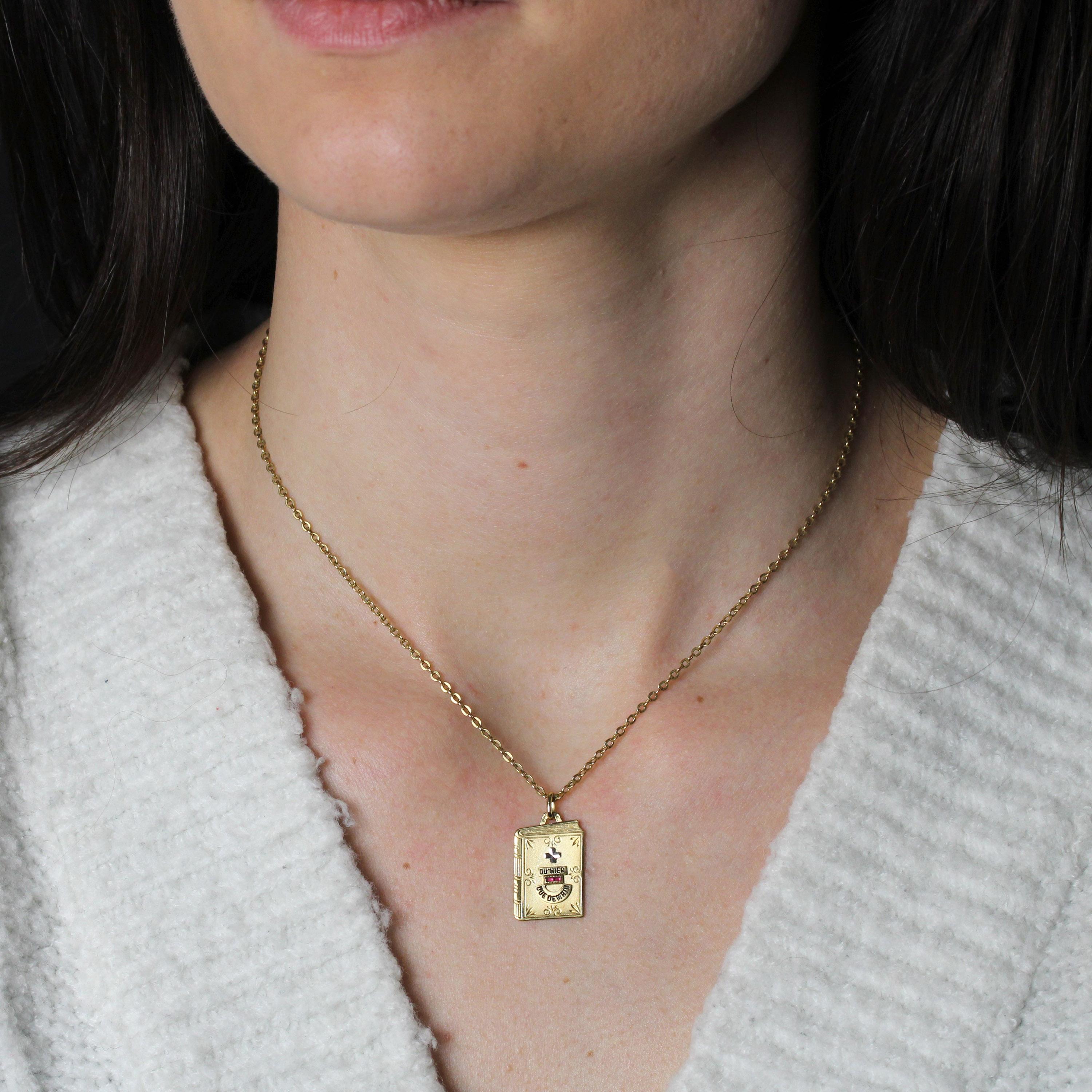 Pendant in 18 karat yellow gold.
This Augis love medal features a matte gold book adorned with Rosemonde Rostand's famous message “+ qu'hier - que demain”, with the “+” in white gold and the “-” adorned with 2 calibrated red gems and the message