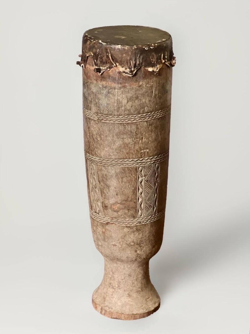 Large, hand-crafted African ceremonial drum sculpted from a single piece of indigenous wood. It features carved geometric motifs and a taut animal hide drum head secured with wooden pegs. The drum has a shapely form with slight asymmetry due to its