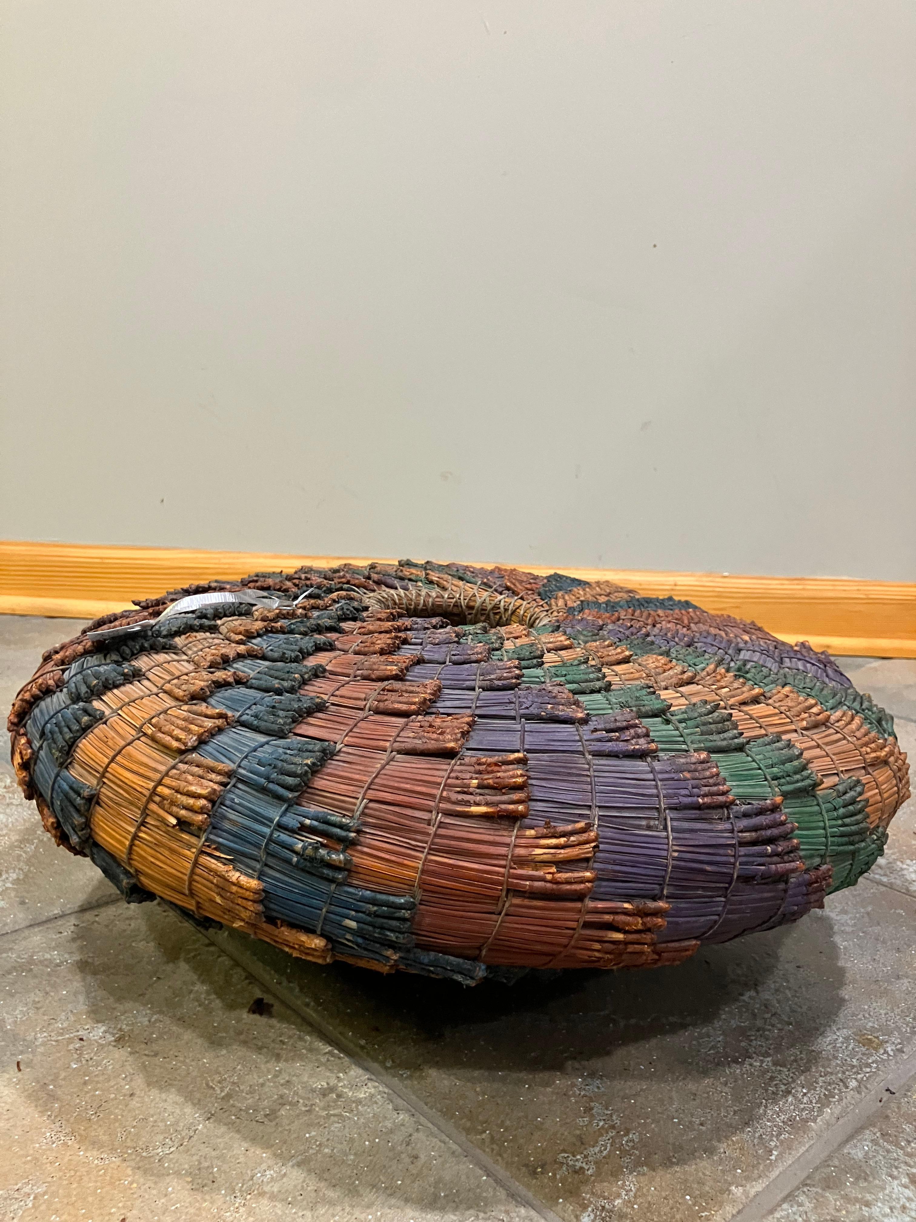 Handwoven African basket made from natural plant fibers, featuring a tightly constructed structure and bold geometric patterning. The surface is composed of dyed reeds in rich tones such as red, blue, orange, purple, green, and black, bound with