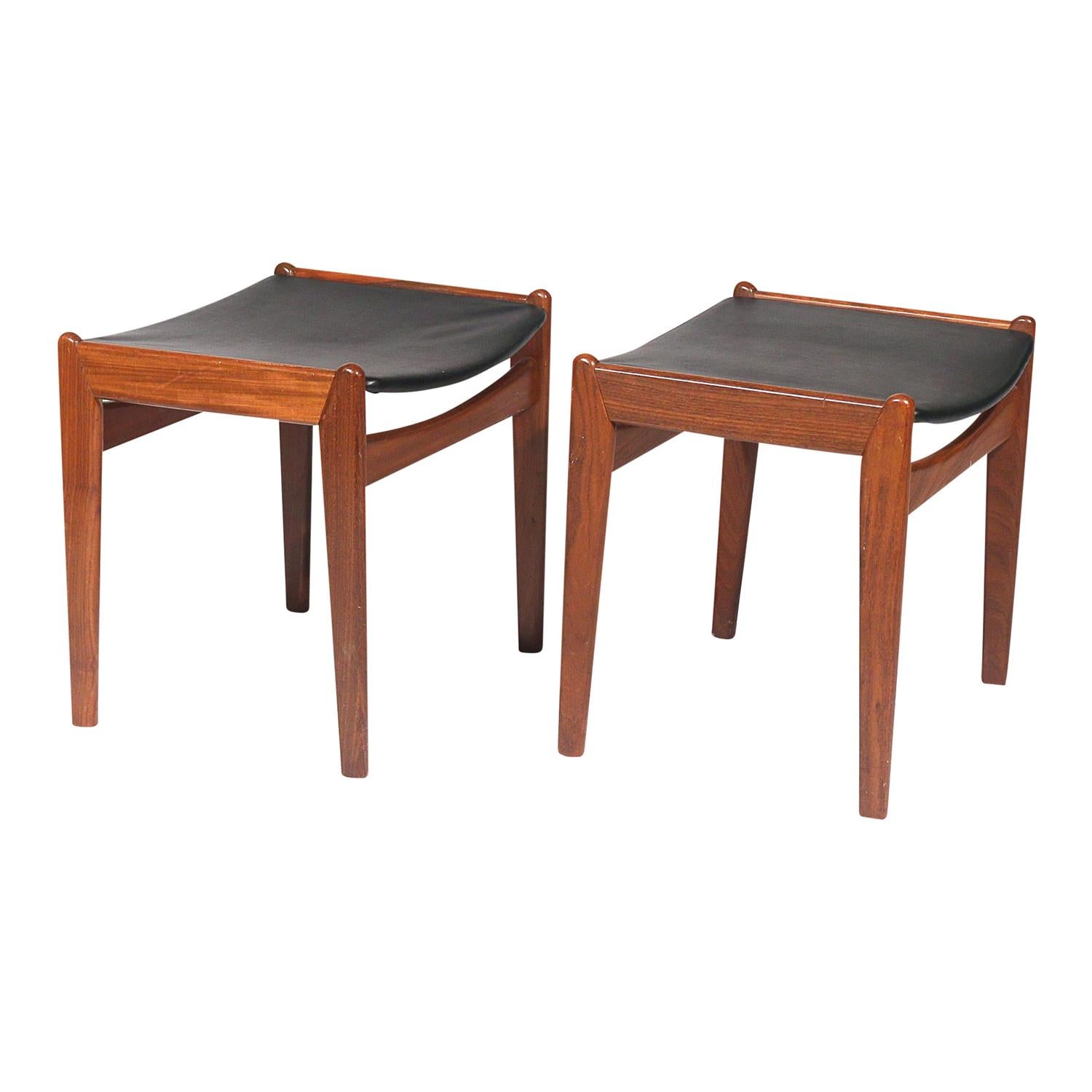 A dark-brown, vintage Mid-Century Modern American pair of footstools made of hand crafted Teakwood in good condition. One of them has still underneath of the seat cover an old label attached of the department store Bloomingdale's. The black vinyl