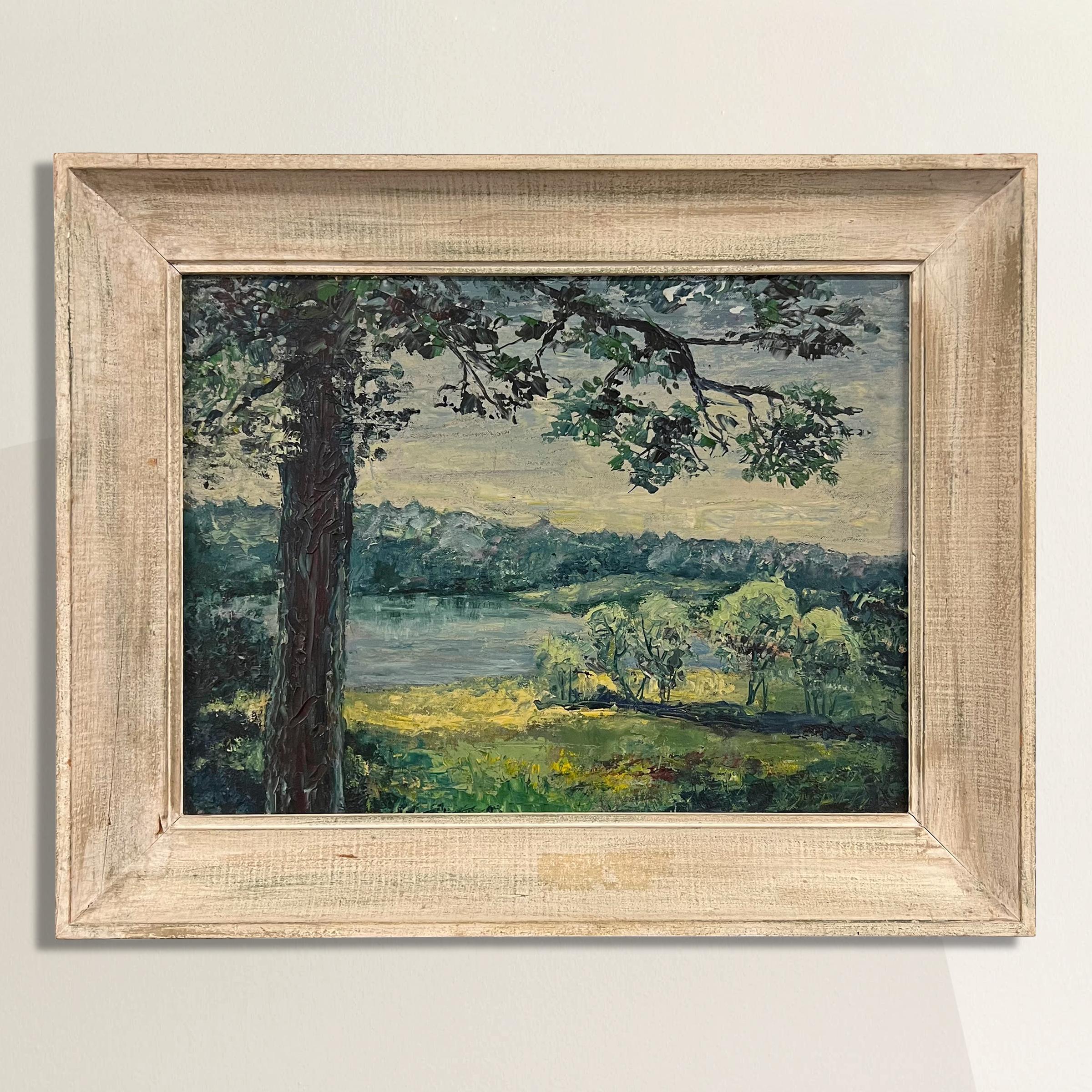 This mid-20th century American plein air painting invites you to a serene lakeside sanctuary.  Framing the composition is a majestic tree in the foreground that stretches towards the heavens and casts gentle shadows over a sunlit meadow. A radiant