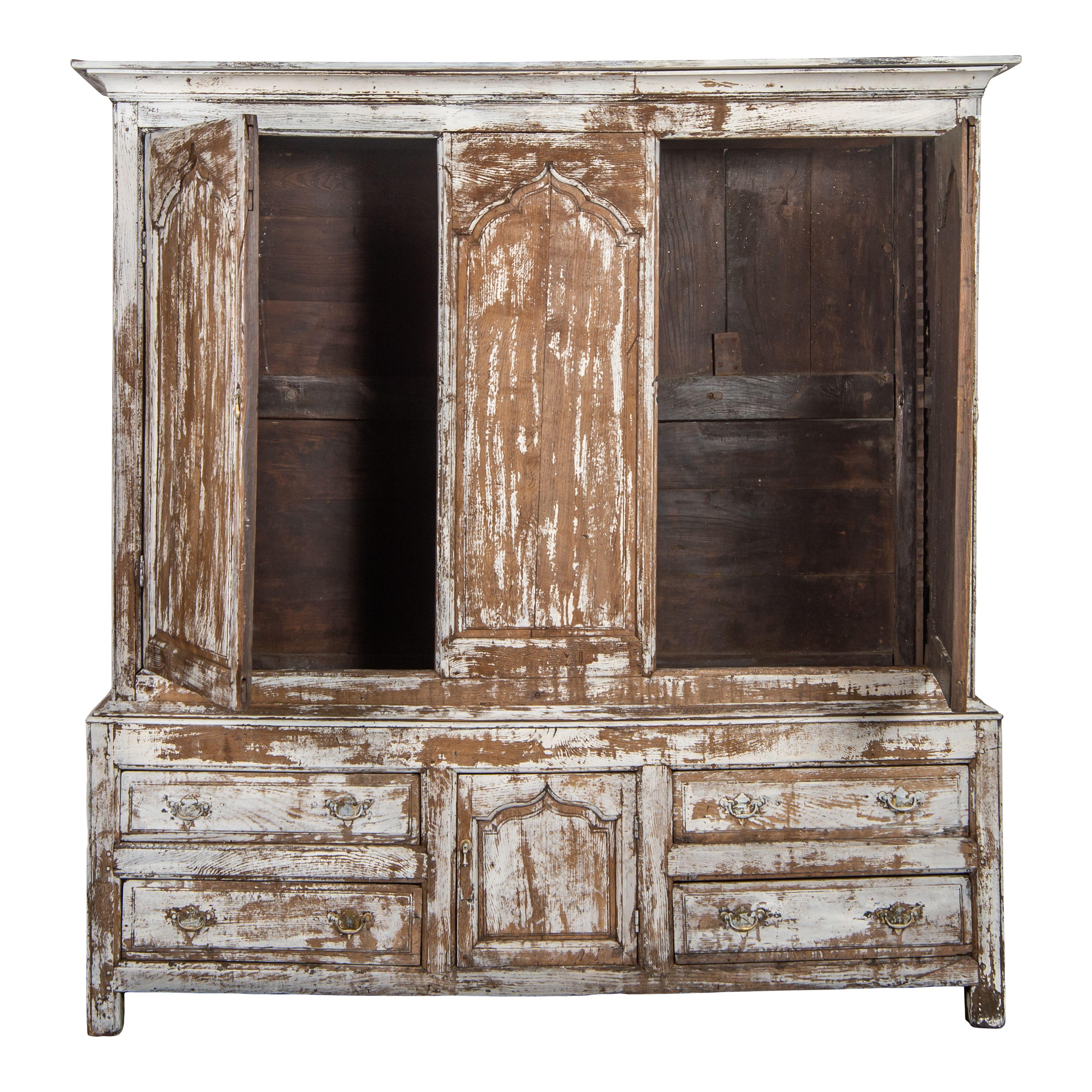 A large vintage wood cabinet from France with chipping white paint; comes in 2 pieces. This large cabinet has a classic wood design, with beautiful carved details framing the doors and drawers and on the moulding. This craftsmanship creates a