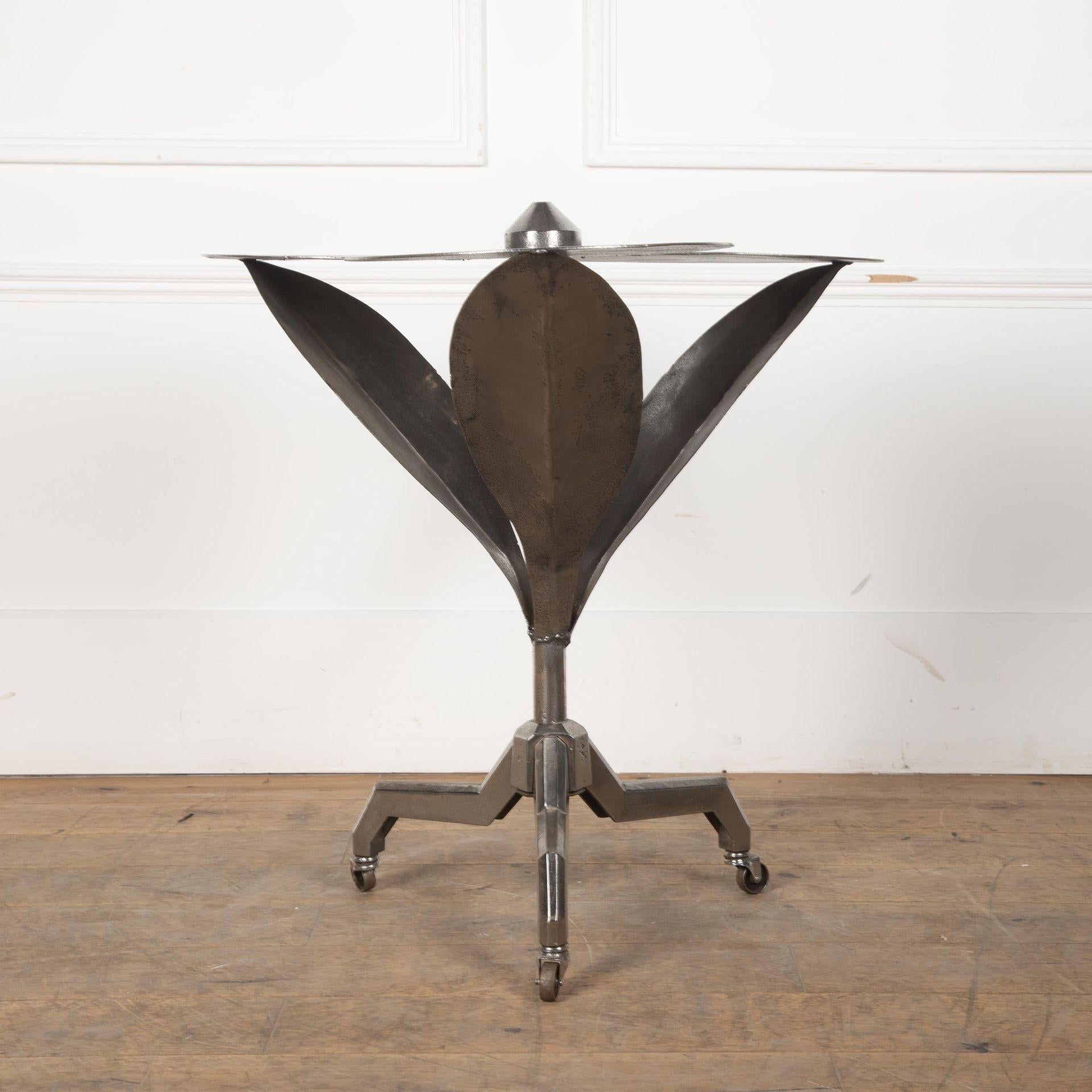 Table d'appoint en acier du début du 20e siècle en forme de fleur stylisée. Trois grands pétales forment le sommet, les branches se raccordant à la colonne tripode, élevée sur trois pieds anguleux munis de roulettes en acier.