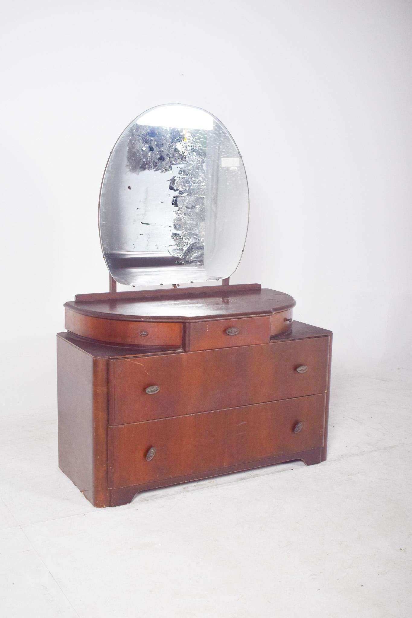 Tocador Art Déco de nogal. Con un imponente espejo circular que descansa sobre una discreta estructura moldeada, refuerza la elegancia modernista del conjunto. La parte superior tiene un recorte curvo que abraza tres cajones pequeños, el central de