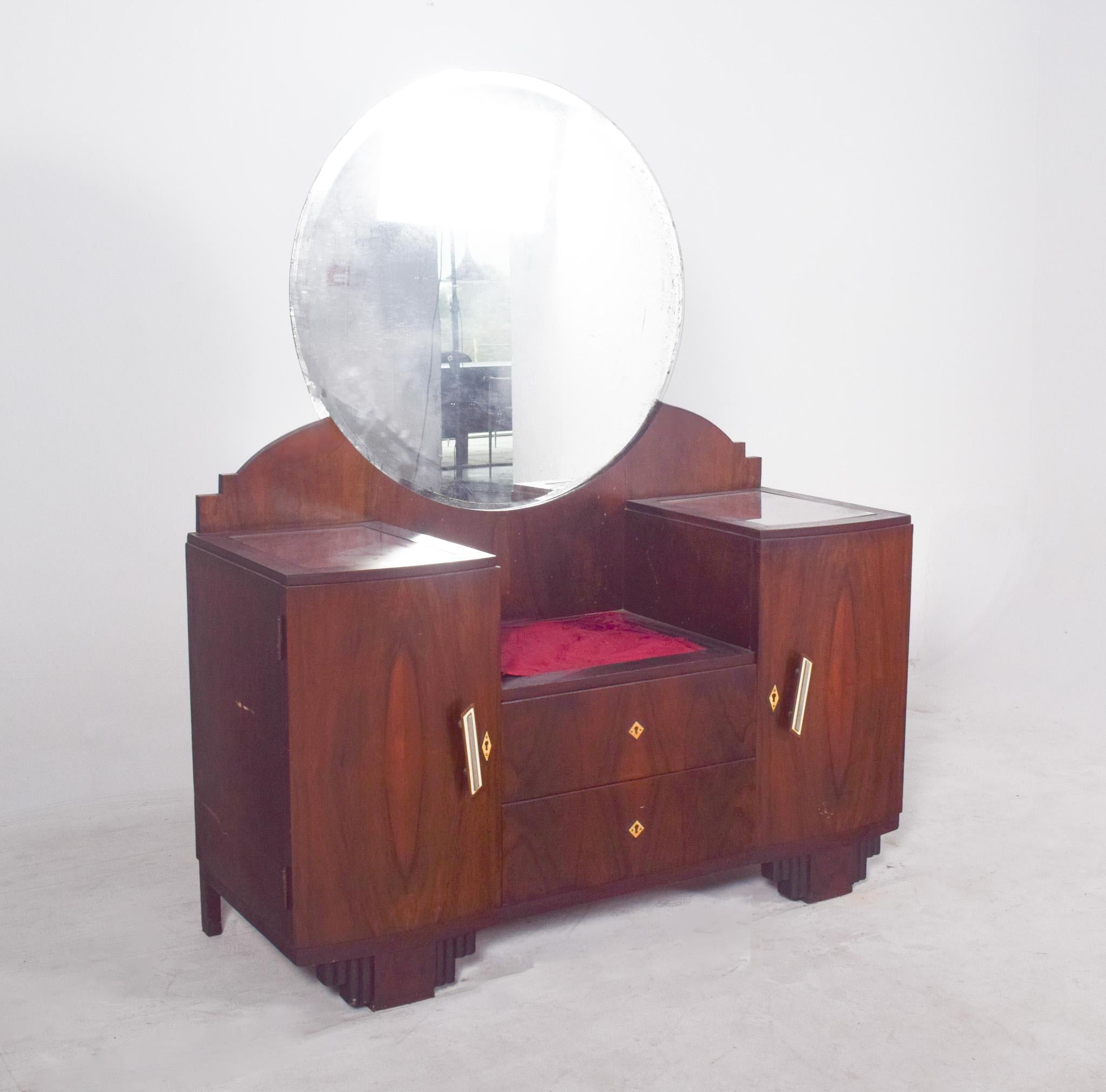 Coiffeuse avec miroir Art Déco. Réalisé en bois noble, il présente des lignes droites et symétriques typiques du style, avec des poignées inclinées en métal doré qui soulignent l'élégance du meuble.
Un grand miroir rond se détache au centre, avec un