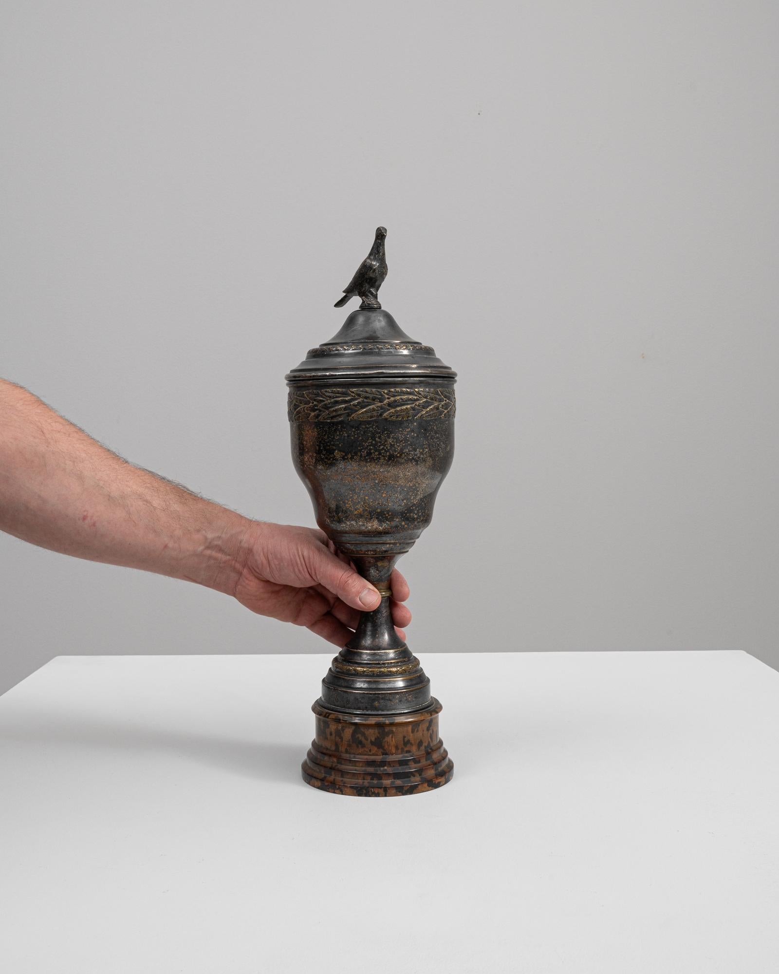 This goblet is a striking piece of 20th-century Belgian artistry, featuring a majestic bird perched atop its lid as if surveying the distance. The dark patina over the brass lends the goblet an air of historical weight and distinction, suggesting