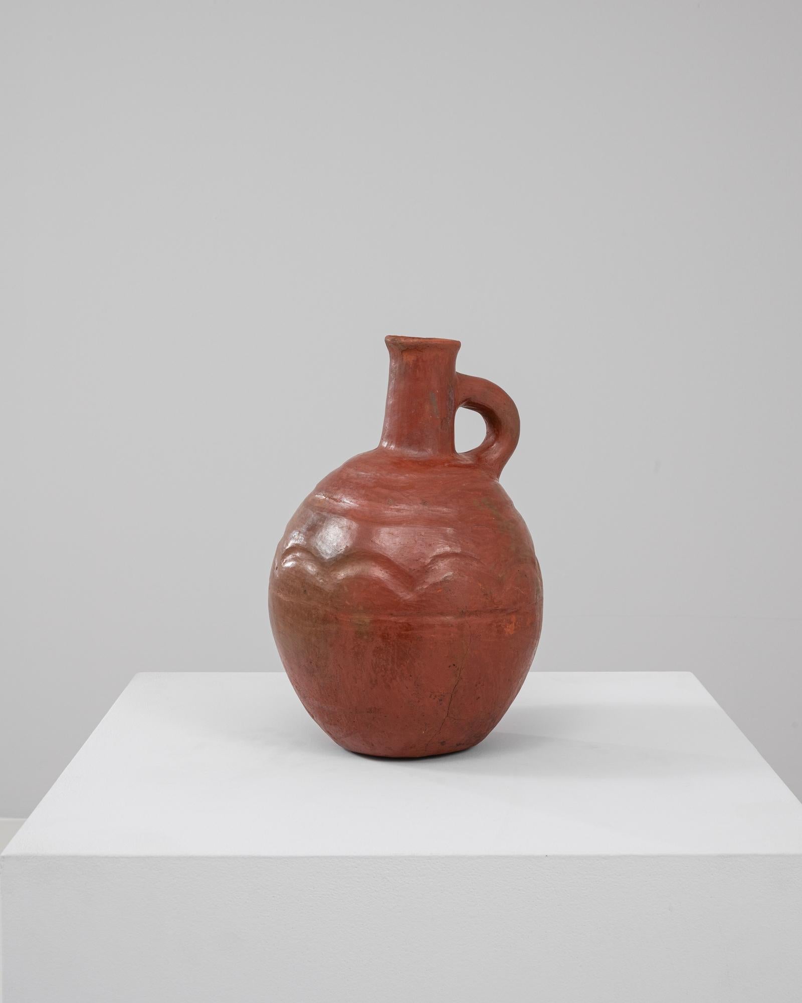 Questa brocca in ceramica belga del XX secolo cattura l'essenza dell'artigianato tradizionale con la sua finitura in argilla rossa e la superficie sottilmente strutturata. Il corpo sferico, abbinato a un beccuccio minimalista e a una maniglia dal