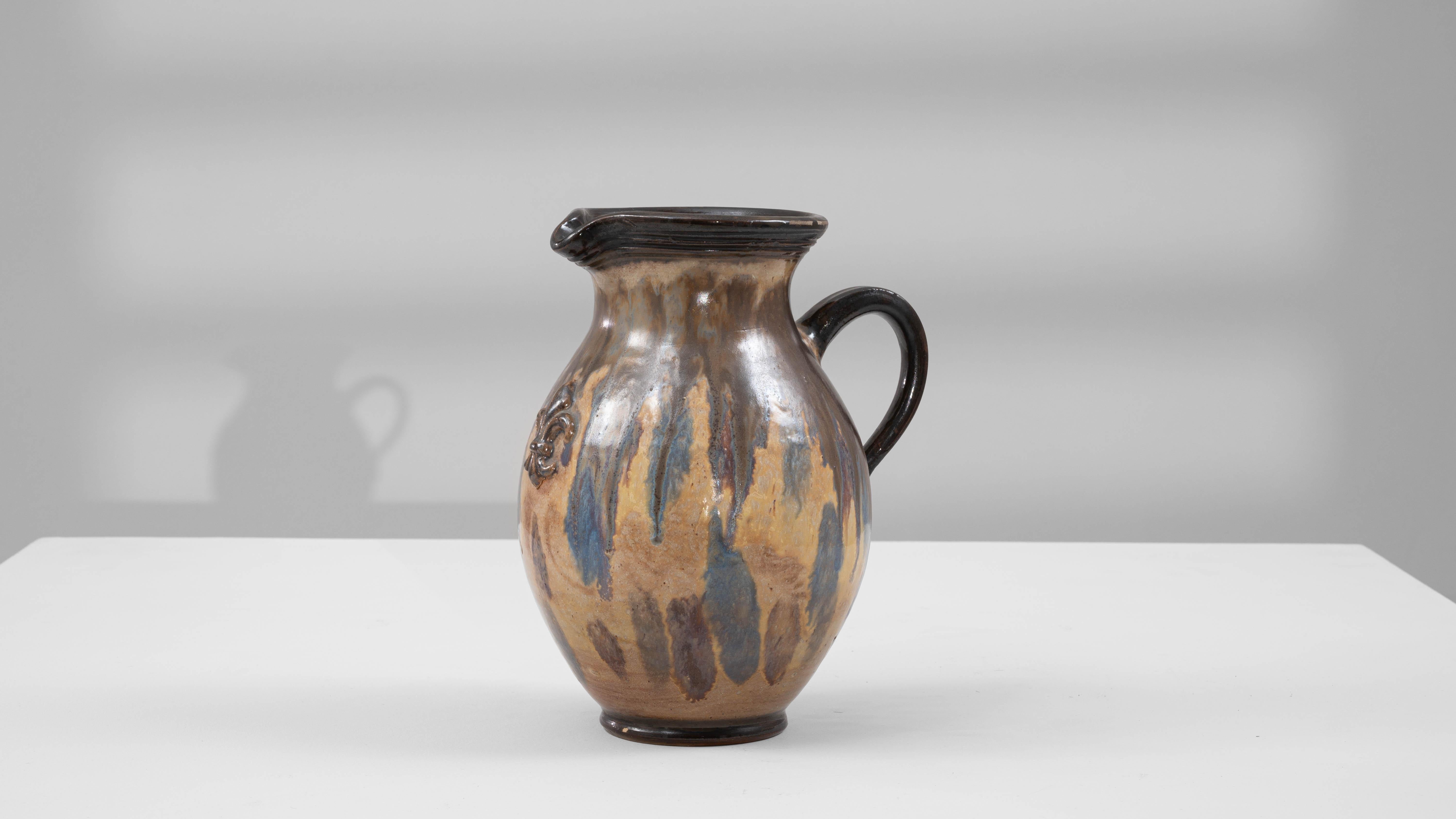 Questa brocca in ceramica belga del XX secolo di G. Losson è una straordinaria fusione di arte e funzionalità. Il suo corpo bulboso mostra un motivo di smalto dinamico con toni terrosi di marrone e beige, accentuati da suggestive gocce blu che