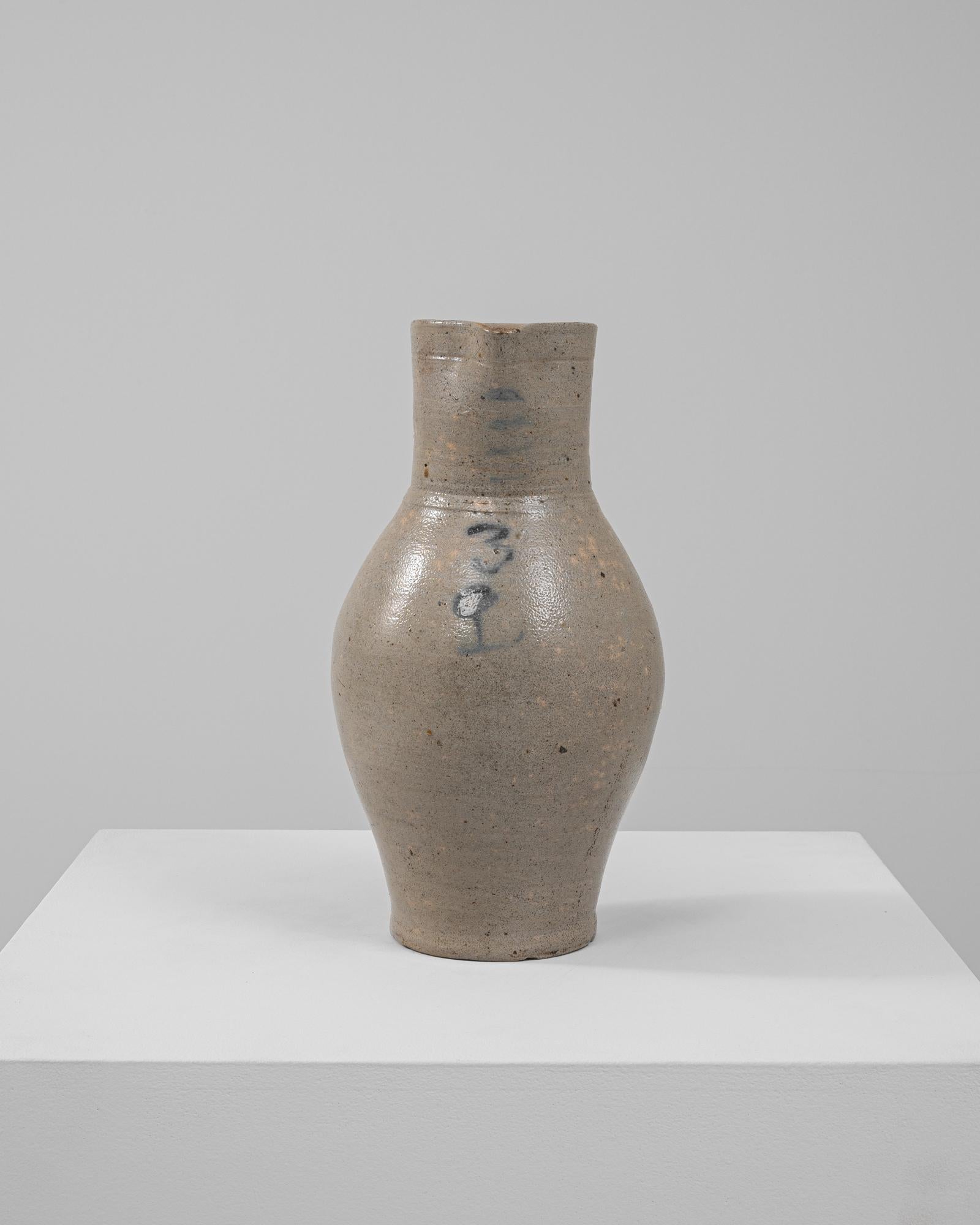 Questa classica brocca in ceramica belga del XX secolo emana un fascino sobrio grazie allo smalto dai toni neutri e al design minimalista. Caratterizzato da una silhouette graziosamente curva e da una robusta maniglia, questo pezzo unisce la