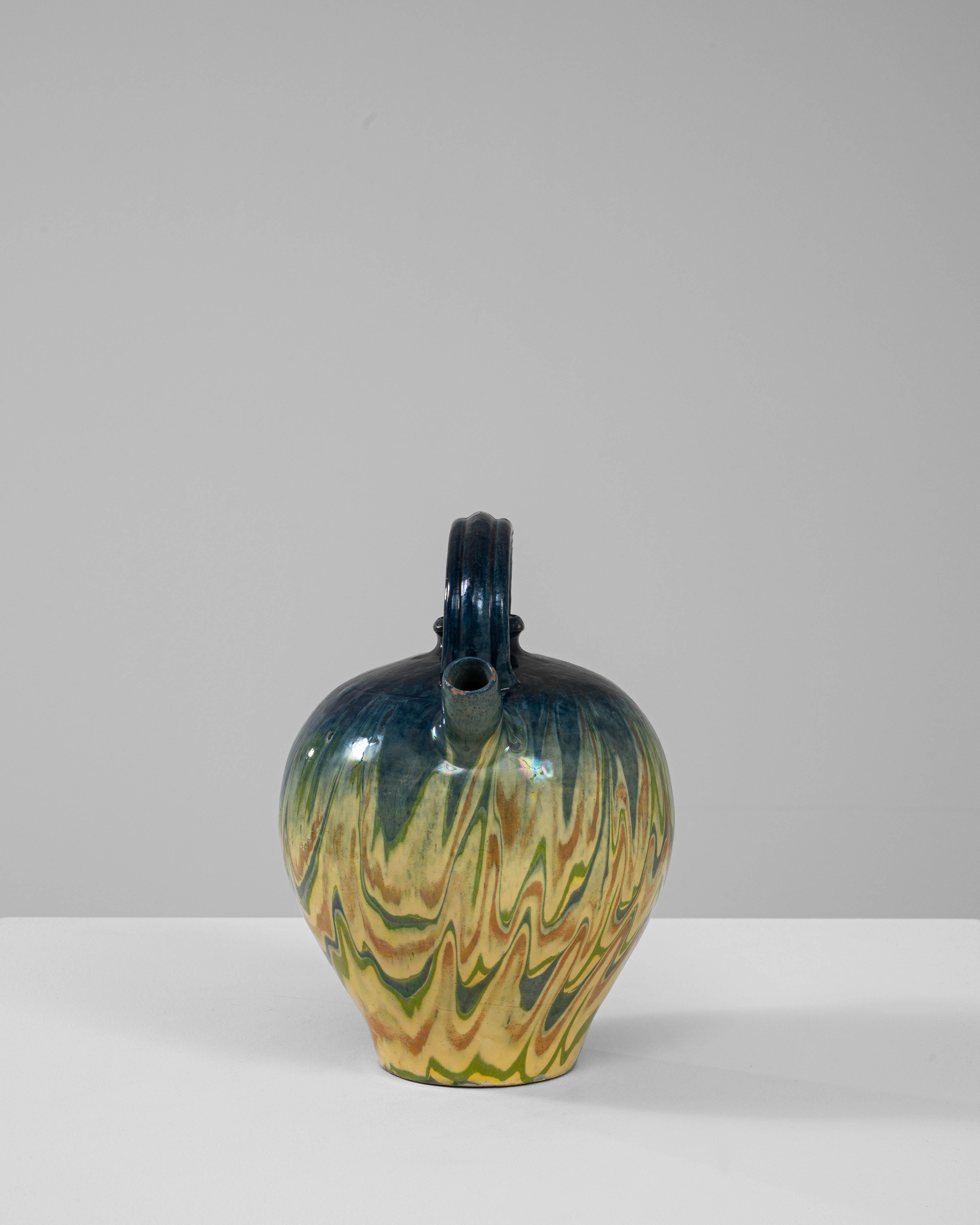 Questa brocca in ceramica belga unica del XX secolo è uno straordinario esempio di artigianato artistico, caratterizzato da un vibrante smalto marmorizzato che mescola gialli, verdi e blu in un dinamico motivo a onde. Il corpo arrotondato della