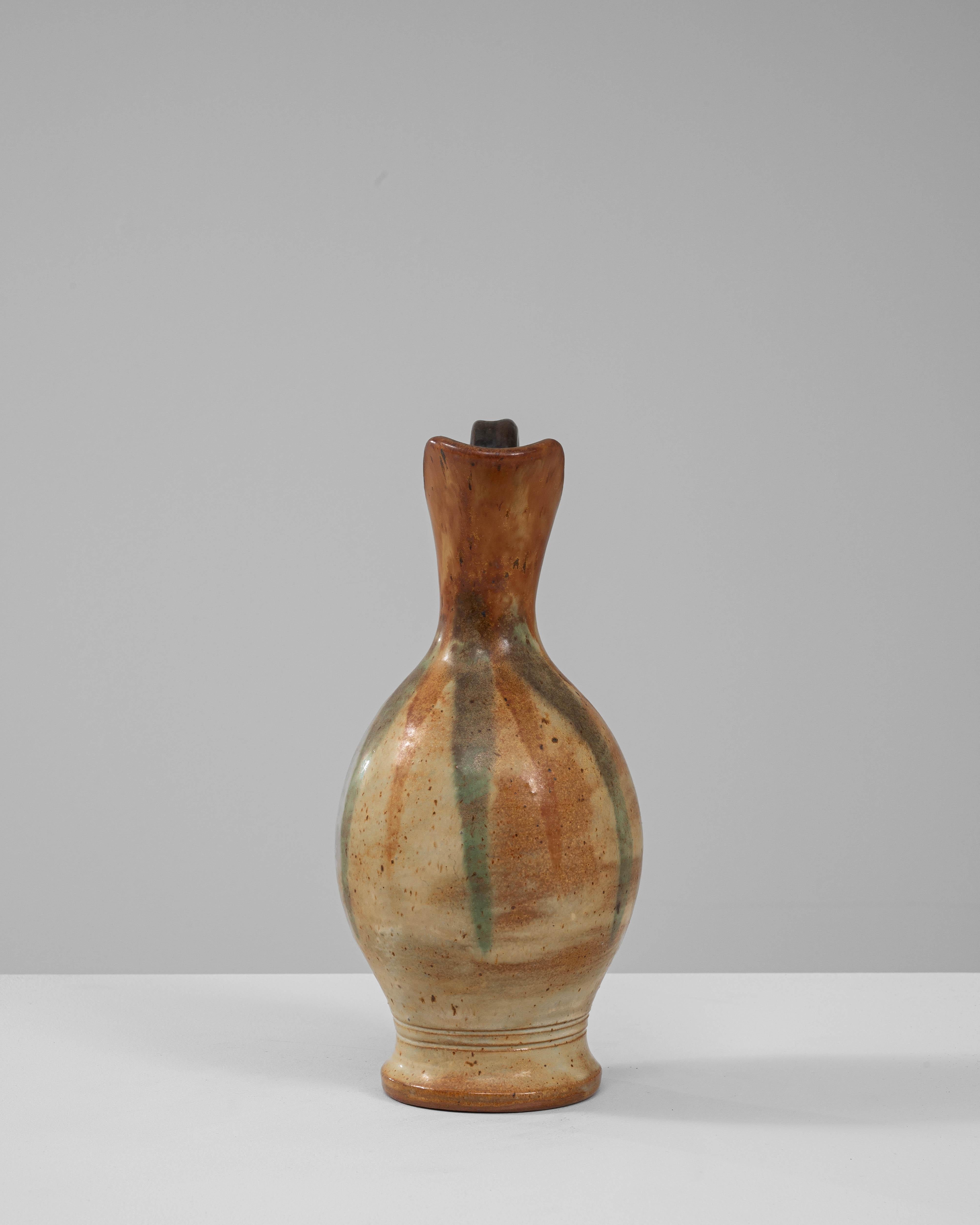 Questa elegante brocca in ceramica belga del XX secolo è caratterizzata da una silhouette senza tempo, con un beccuccio graziosamente curvo e un manico robusto e arcuato. Lo smalto caldo e terroso passa dal beige sabbioso ai toni sottili del verde e