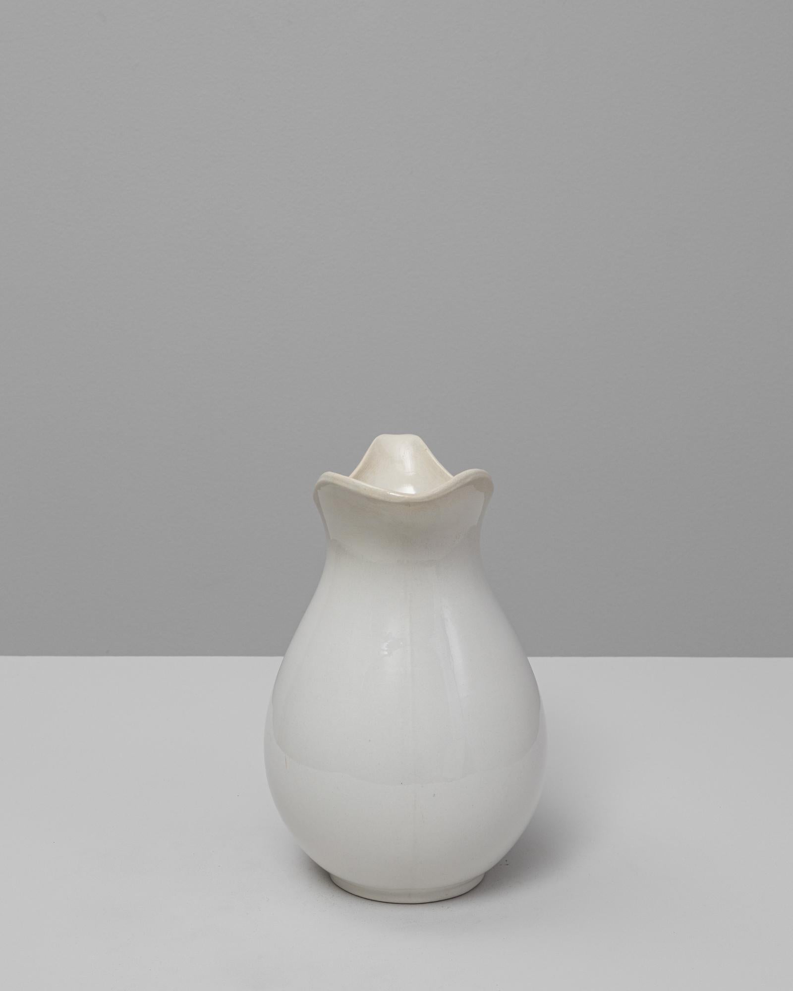Pichet en céramique belge du 20e siècle. Cet élégant pichet en céramique émaillée blanche présente une silhouette doucement incurvée avec un large bord festonné et une poignée gracieuse. Sa forme simple et son émail subtilement craquelé lui