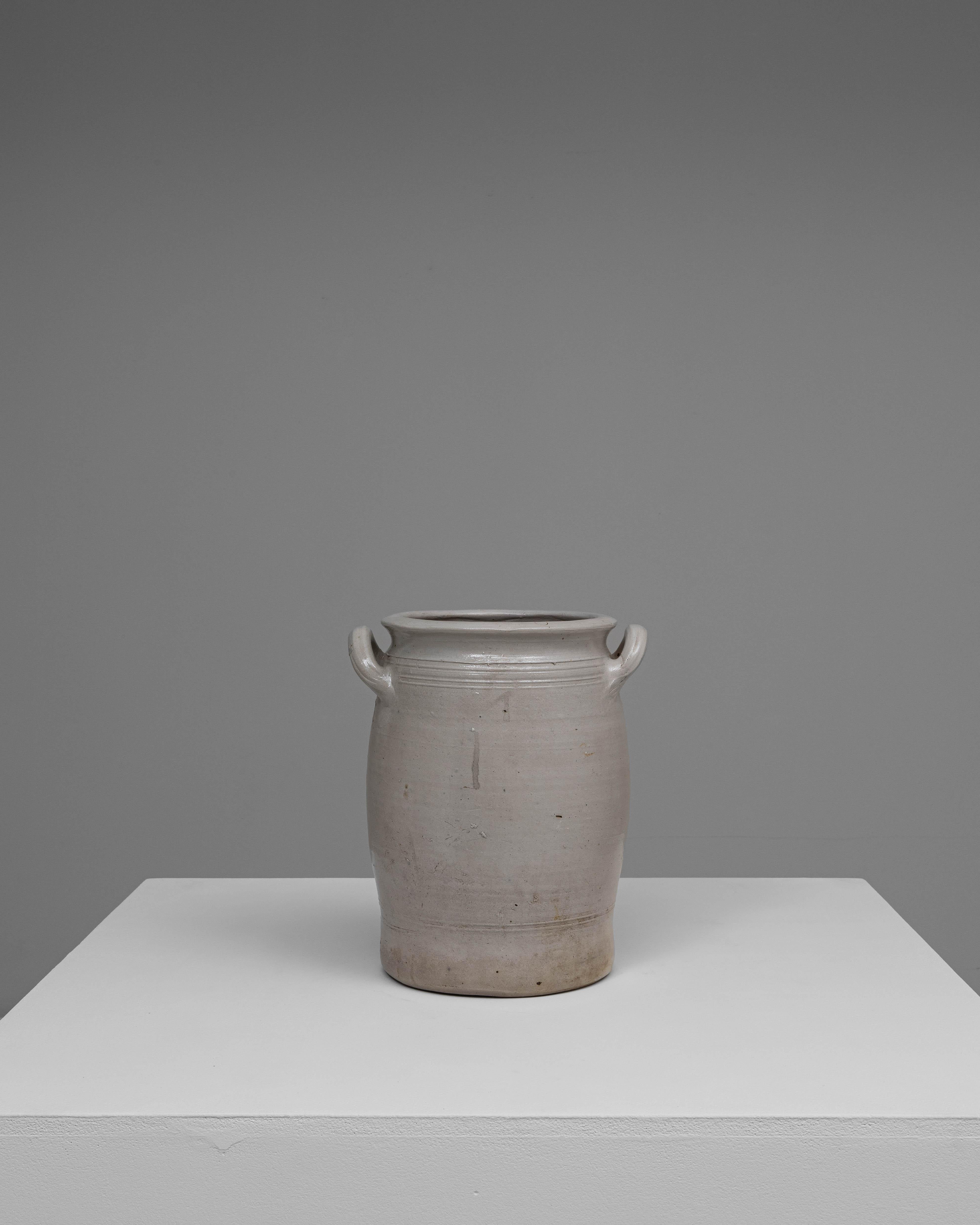 Ce pot en céramique belge du XXe siècle présente une forme simple et utilitaire, rehaussée par un travail artisanal subtil et une surface doucement vieillie. Le corps cylindrique se rétrécit doucement vers la base, tandis qu'une large embouchure au