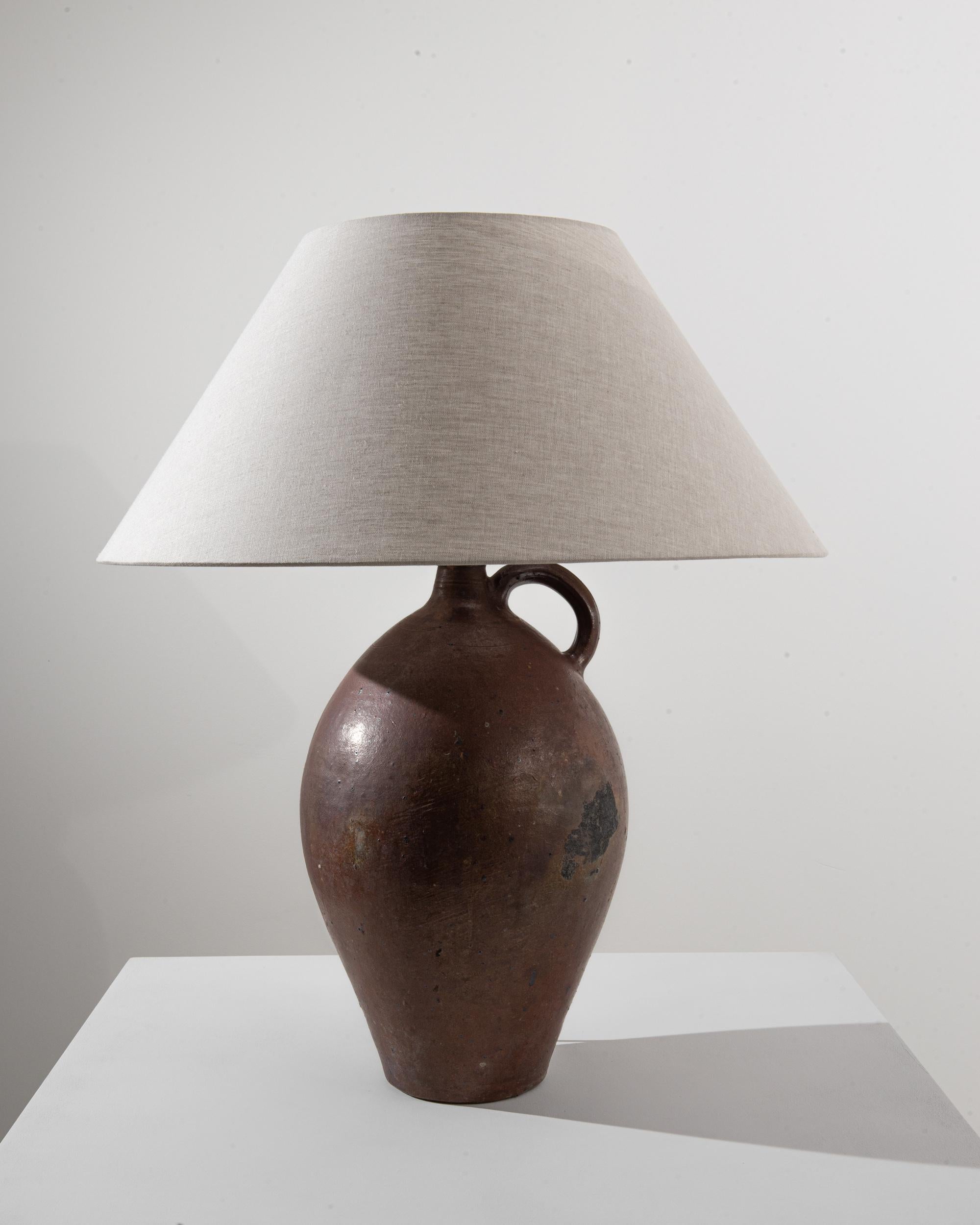 A 20th century ceramic table lamp produced in Belgium, this antique pottery piece is magnetized with haptic force. Made of a large repurposed pitcher with a handle and a chiffon shade, this warm light displays a full body in a lush chocolate tone,