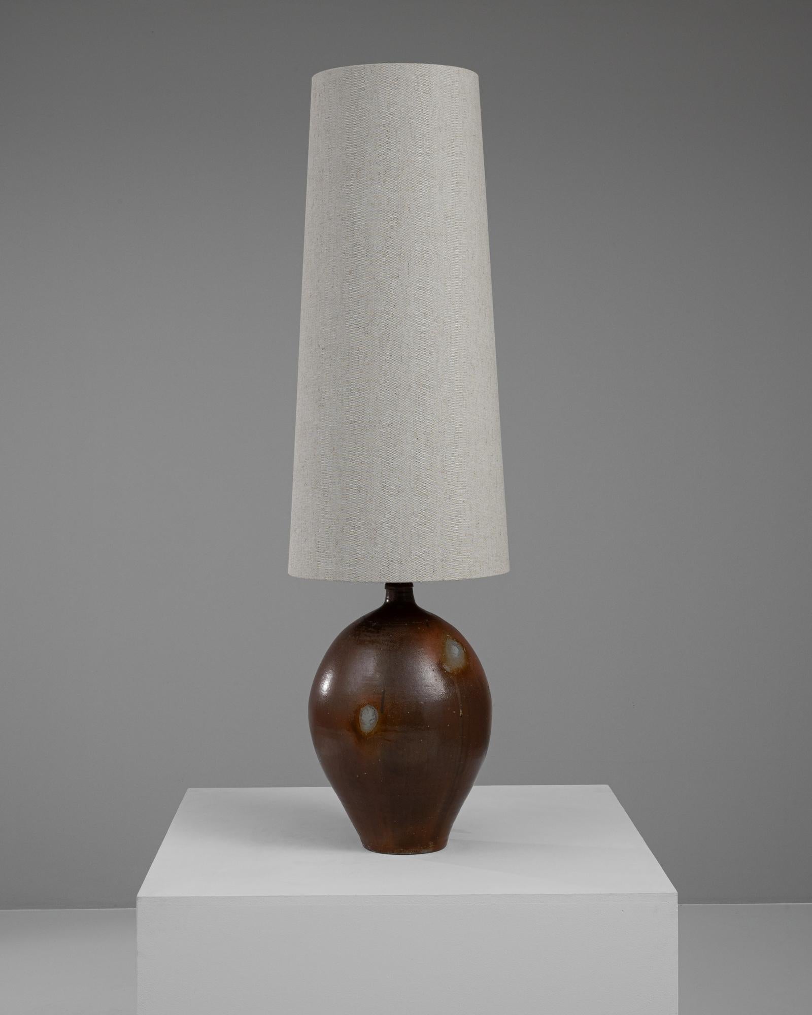 This 20th-century Belgian ceramic table lamp encapsulates a blend of simplicity and rustic charm, making it an ideal addition to any home decor. The base is crafted from high-quality ceramic, featuring a rich, glossy brown finish with natural,